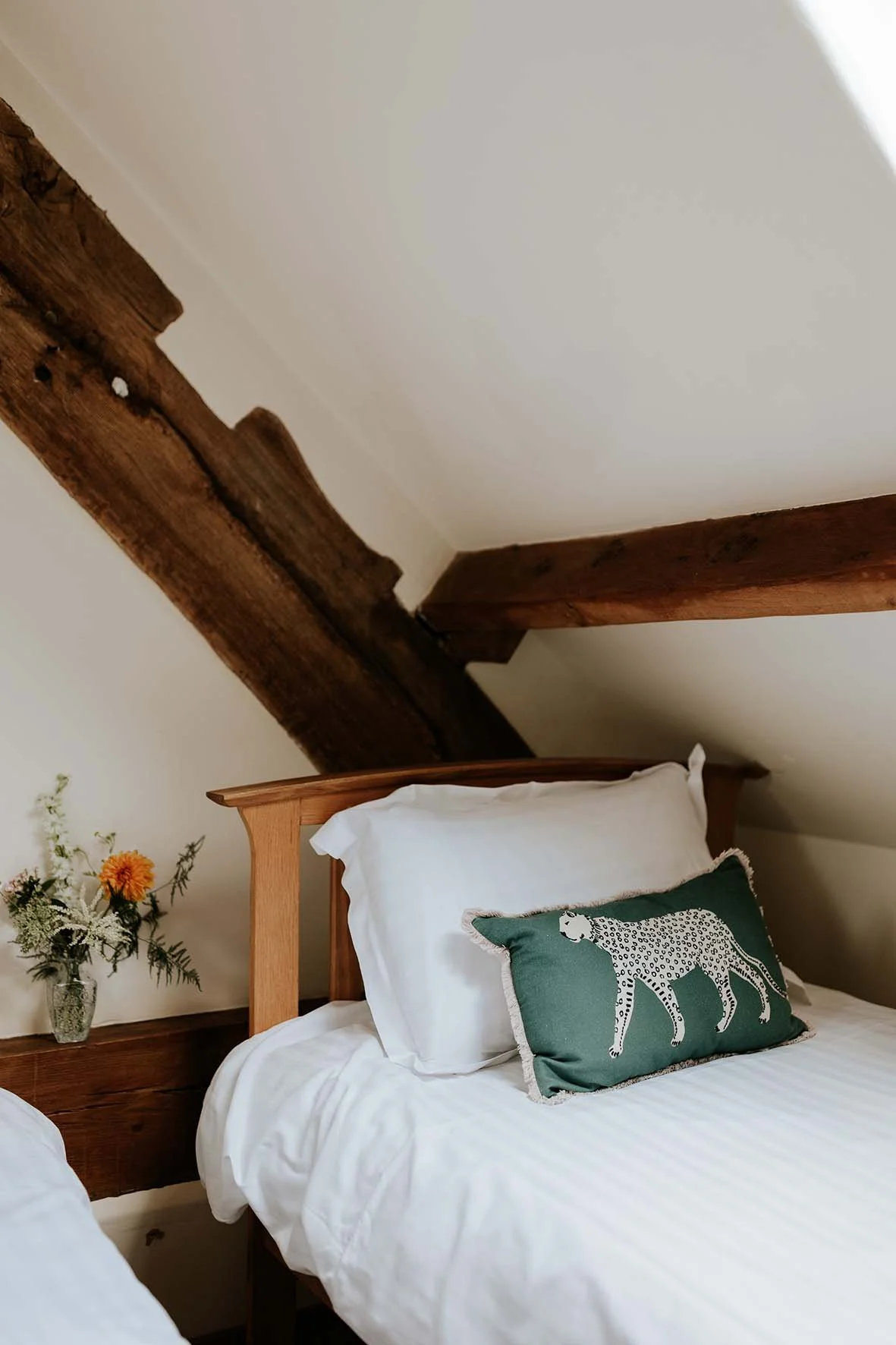 A bedroom corner with a bed adorned with white bedding and a decorative pillow featuring a leopard illustration, a small side table with a flower arrangement, and exposed wooden beams on the ceiling.