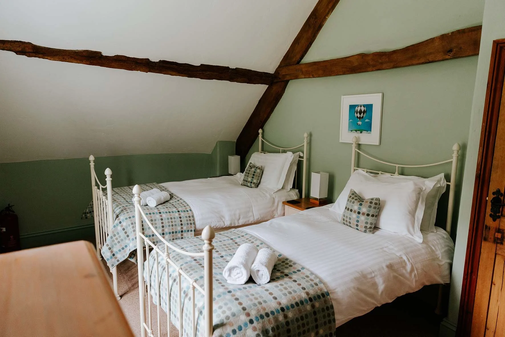 Two single beds with white metal frames and polka dot bedspreads, towels, and pillows in a bedroom with sloped ceiling, green walls, and exposed wooden beams.