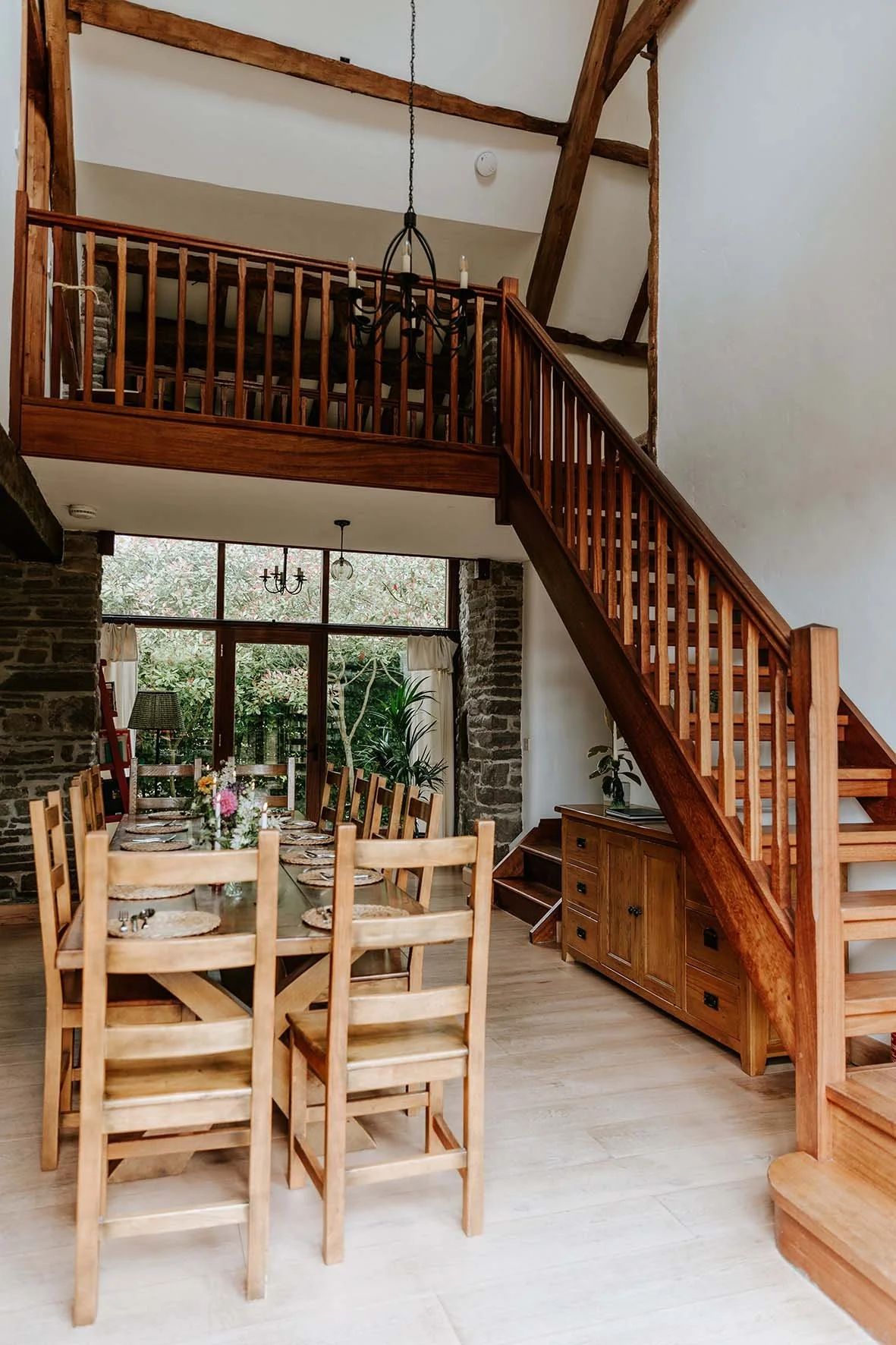 A dining area with a long wooden table set for a meal, surrounded by matching wooden chairs. The room features a stone accent wall and large windows overlooking a garden. There is a wooden staircase leading to a loft area, and chandeliers hang from t