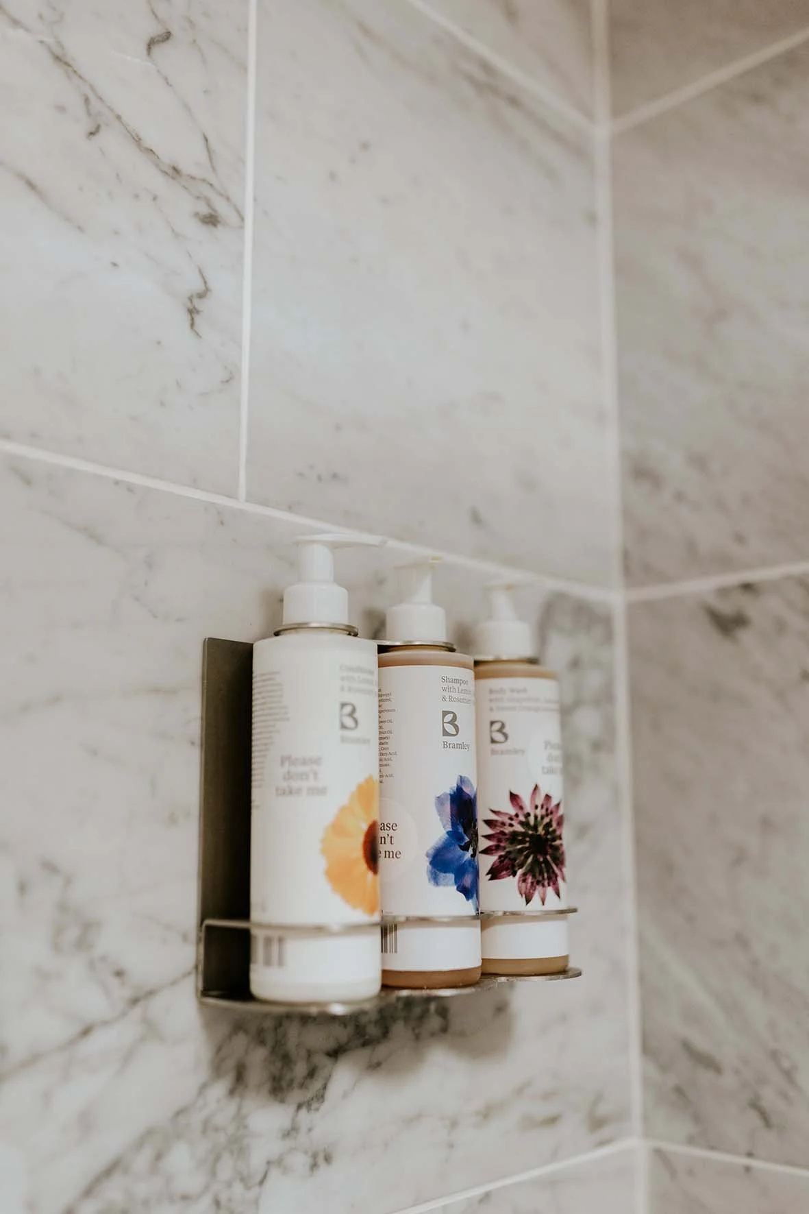 Three bottles of shampoo with floral labels, mounted on a metal holder on a marble tiled wall.