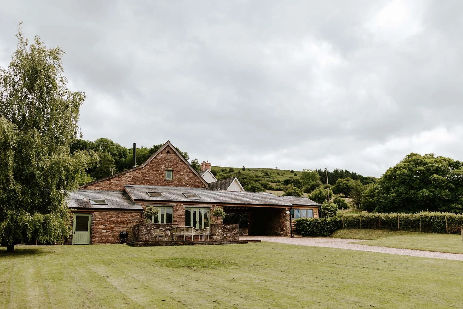 A rural house made of brick with a large lawn and surrounded by trees and hills under cloudy sky.