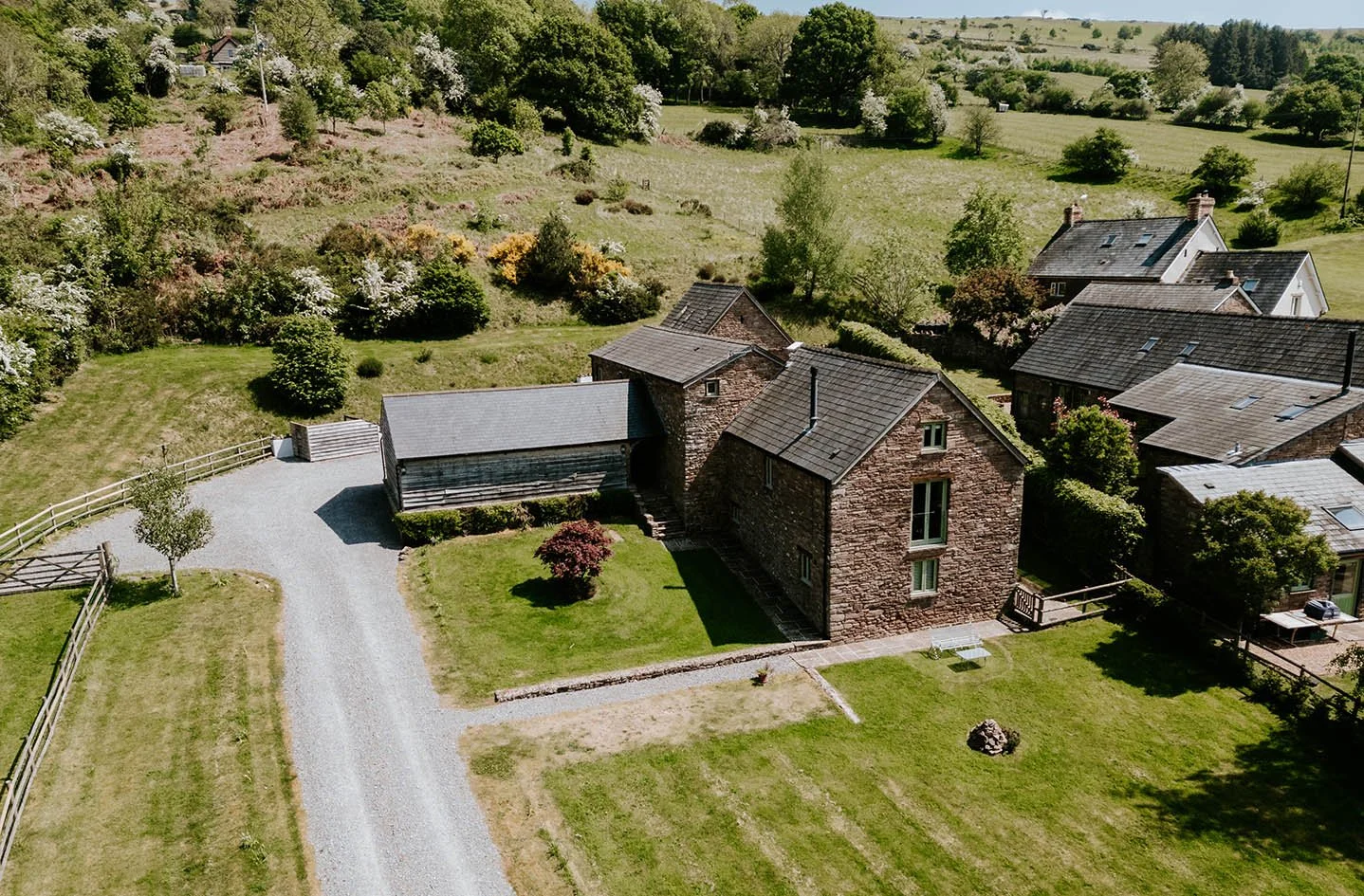 Aerial view of a rural property with a main stone house, a wooden barn, and surrounding green lawns, trees, and neighboring houses on a sunny day.