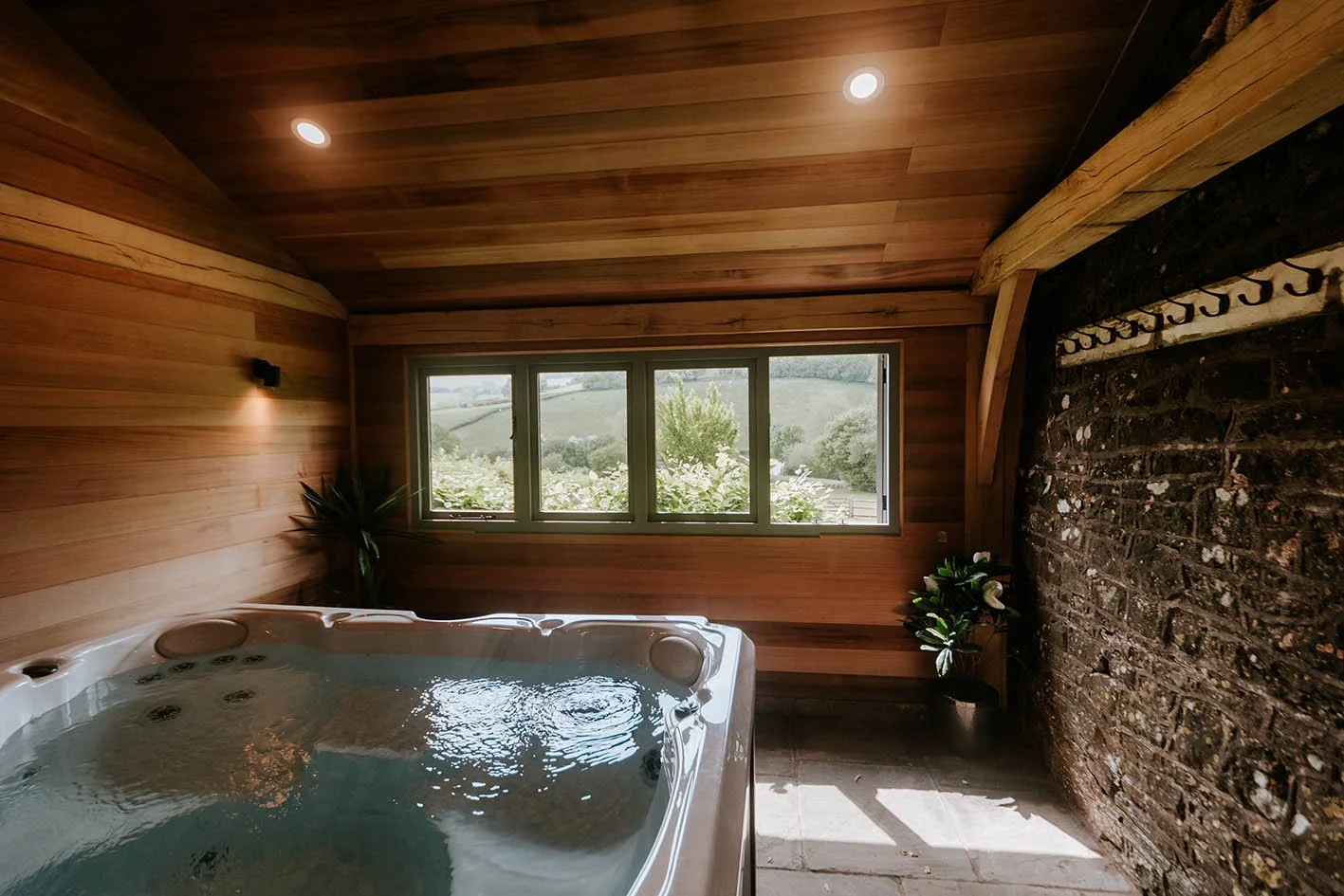 Indoor hot tub with water on a wooden deck, large window overlooking a countryside with rolling hills, potted plants, and a brick wall on the right.