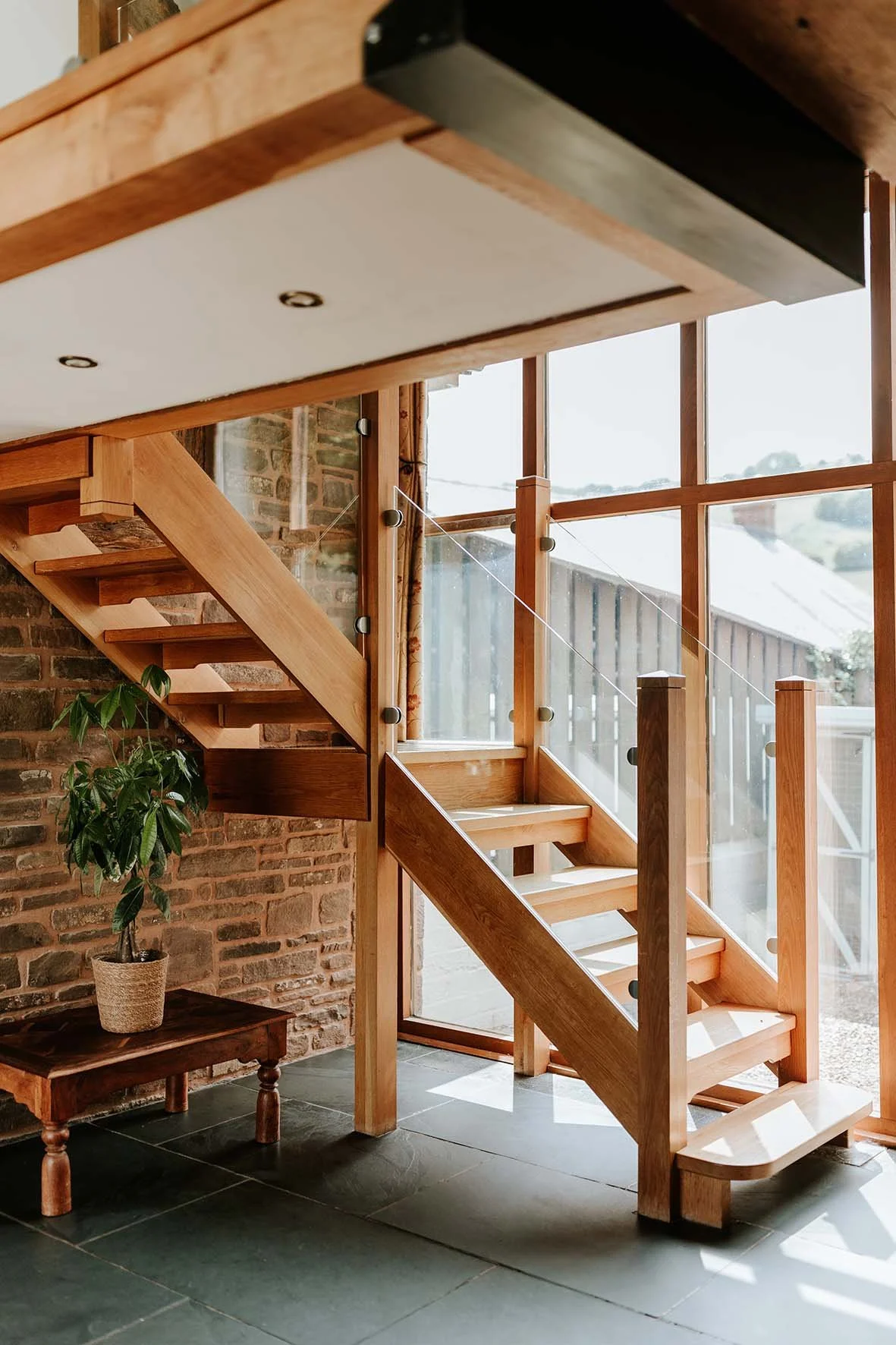 Interior view of a wooden staircase next to large windows with a wooden railing, brick wall, small wooden table with a potted plant, and green tiled floor.