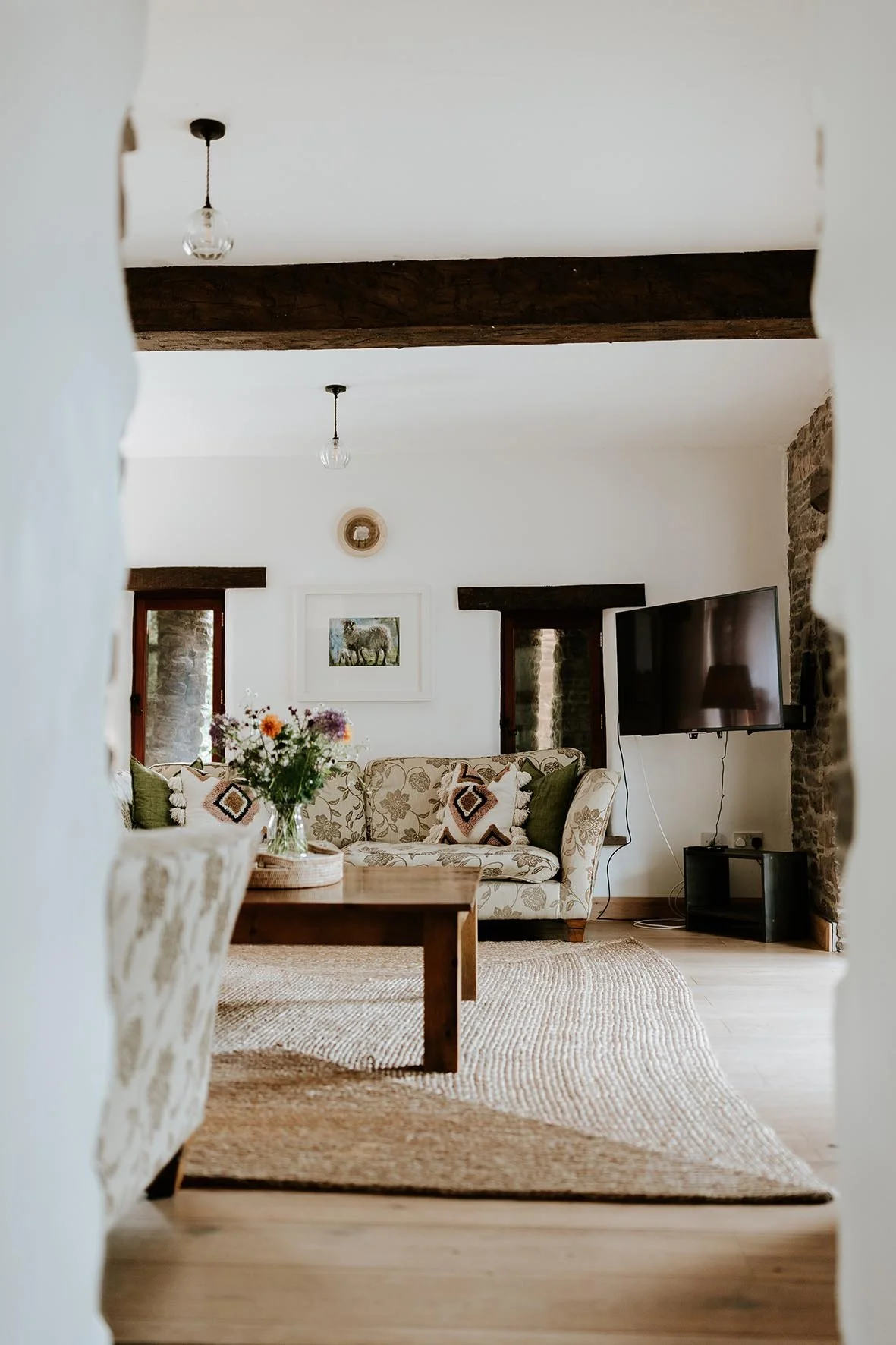 Cozy living room with a floral patterned sofa, a wooden coffee table with a vase of flowers, a wall-mounted flat-screen TV, stone fireplace, and rustic wooden ceiling beams.