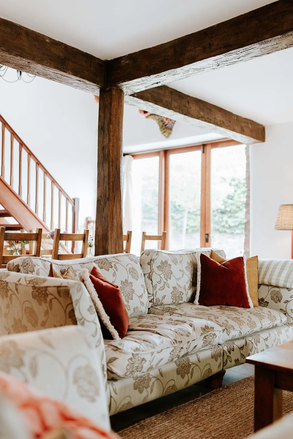 Living room with cream floral-patterned sofa, red and brown pillows, wooden beams, large window with white curtains, and staircase. A cat is partially visible on the ceiling.