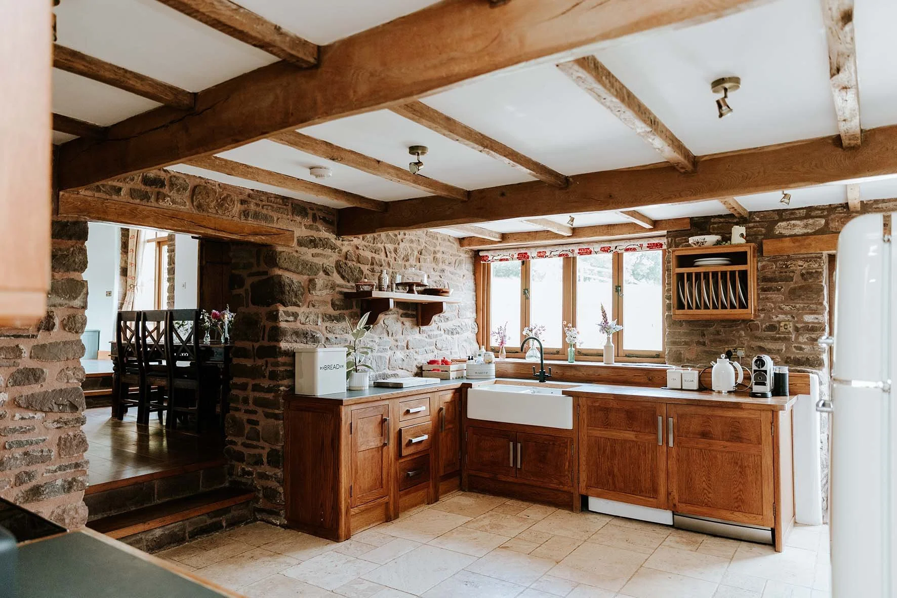 Rustic kitchen with wooden cabinets, brick walls, and large window with floral valance, overlooking a dining area.