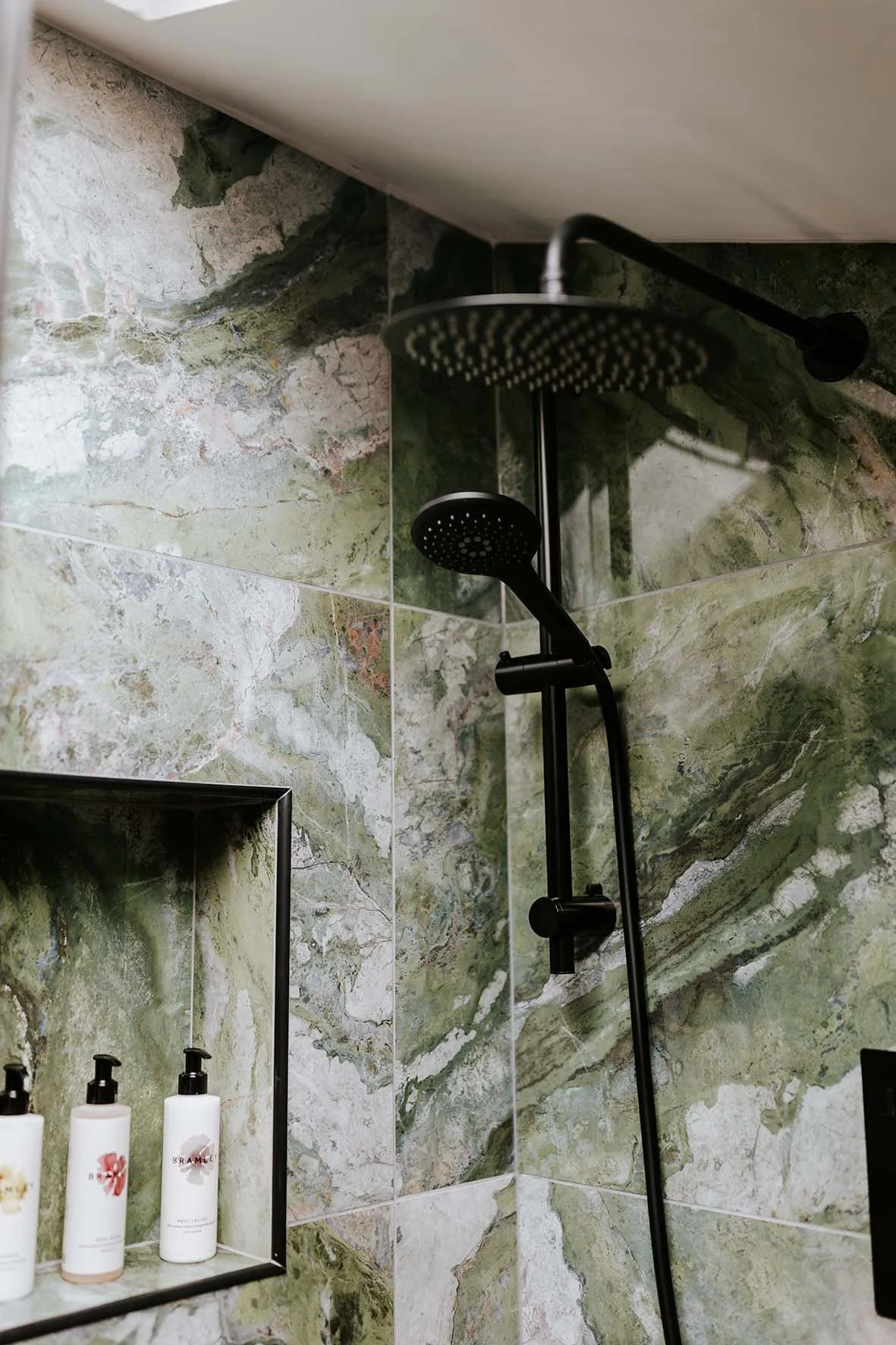 A bathroom shower area with green and beige marble tile walls, featuring a black shower head and a built-in shelf with three bottles of soap or lotion.