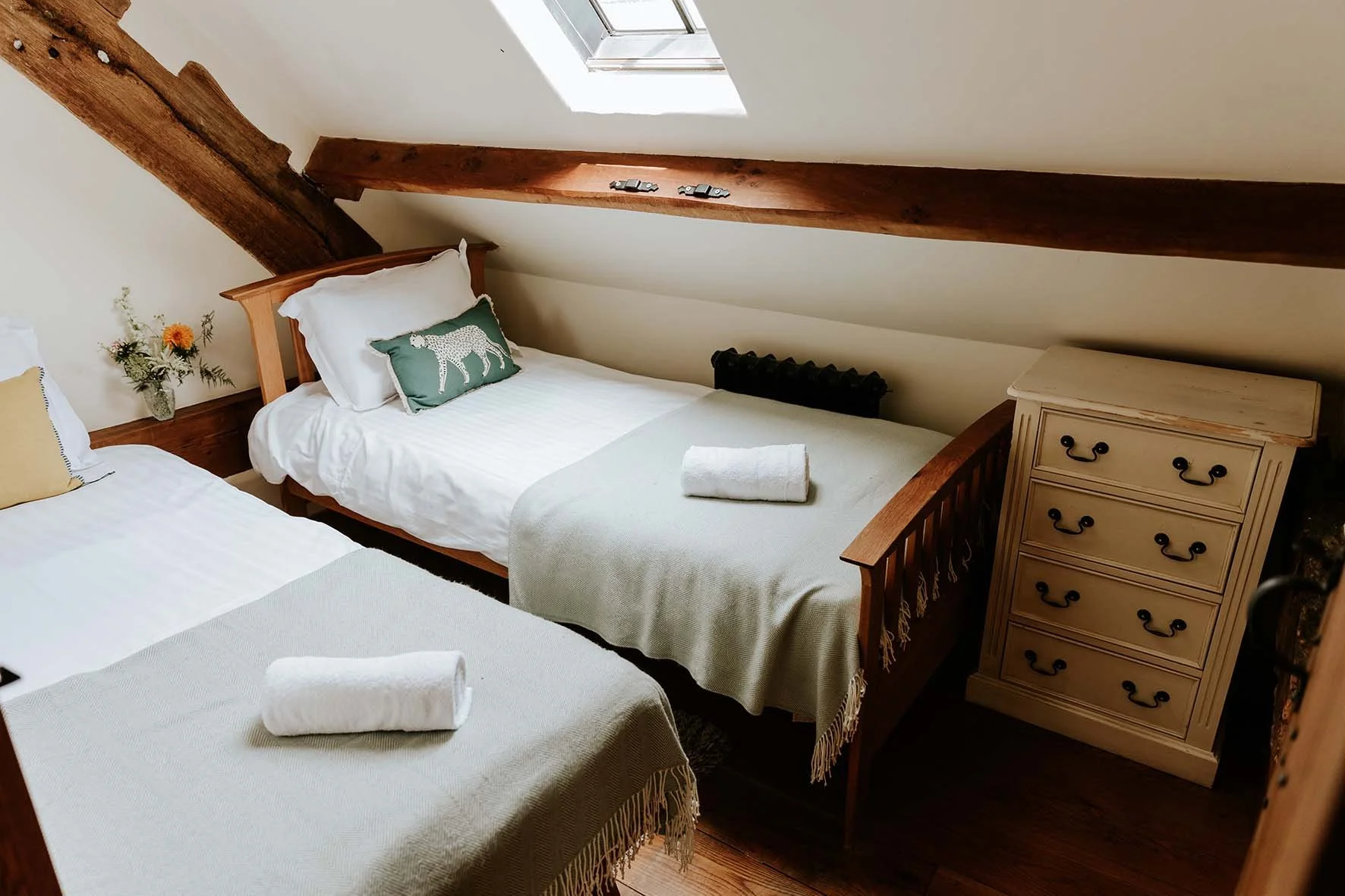 Bedroom with two twin beds, wooden headboard and frames, white bedding, rolled towels, a nightstand with black handles, a small vase with flowers, a skylight, and wooden beams on the ceiling.