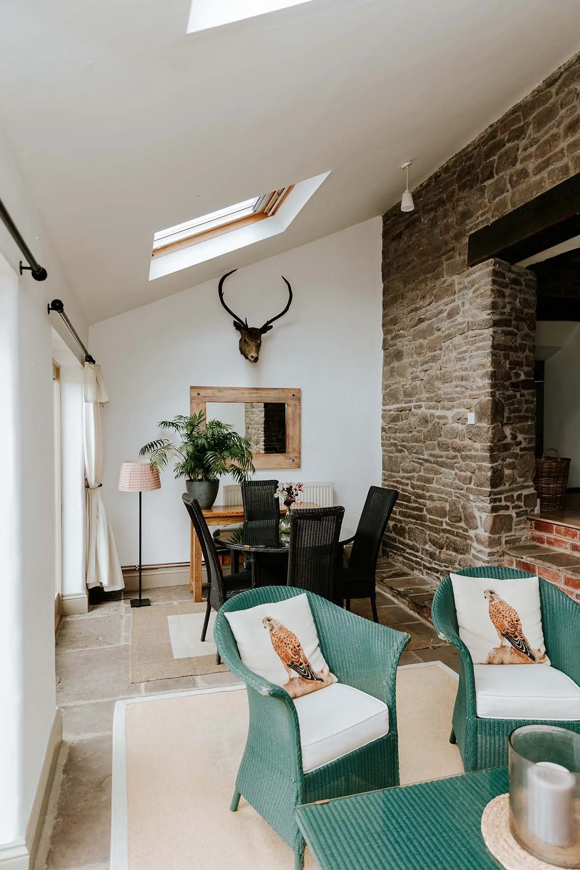 Living room with a high, angled ceiling, skylight, mounted deer head, dining table with chairs, green armchairs with bird pillows, brick wall, and a mirror.