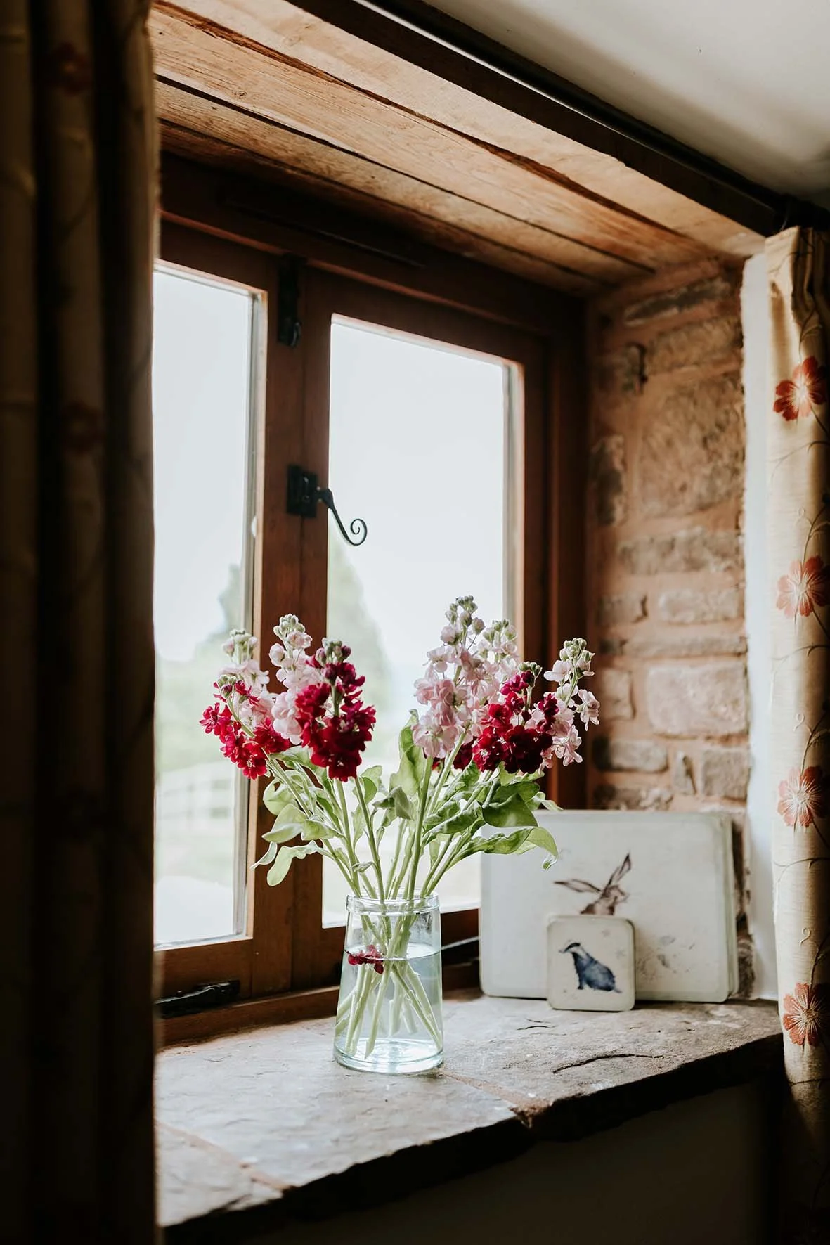 A glass vase with pink and white flowers on a windowsill in a rustic room with brick walls and floral curtains.