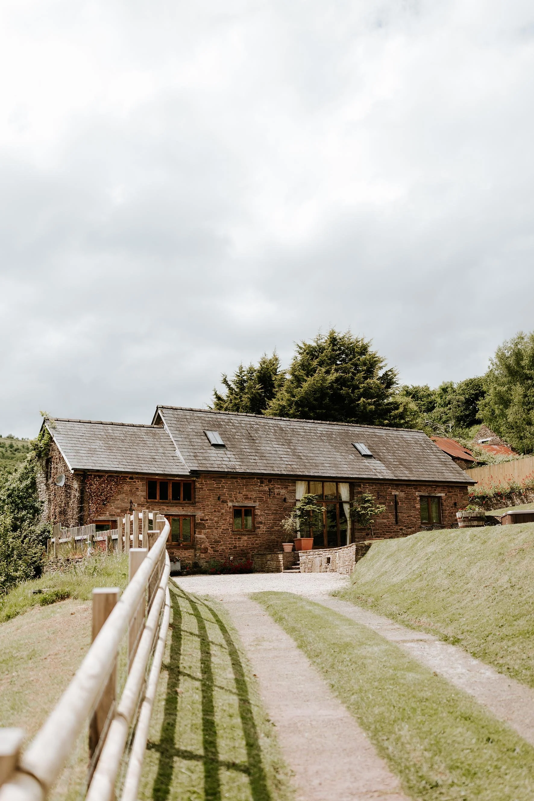 A rustic brick house with a shingled roof, surrounded by greenery and a gravel driveway, under a cloudy sky.