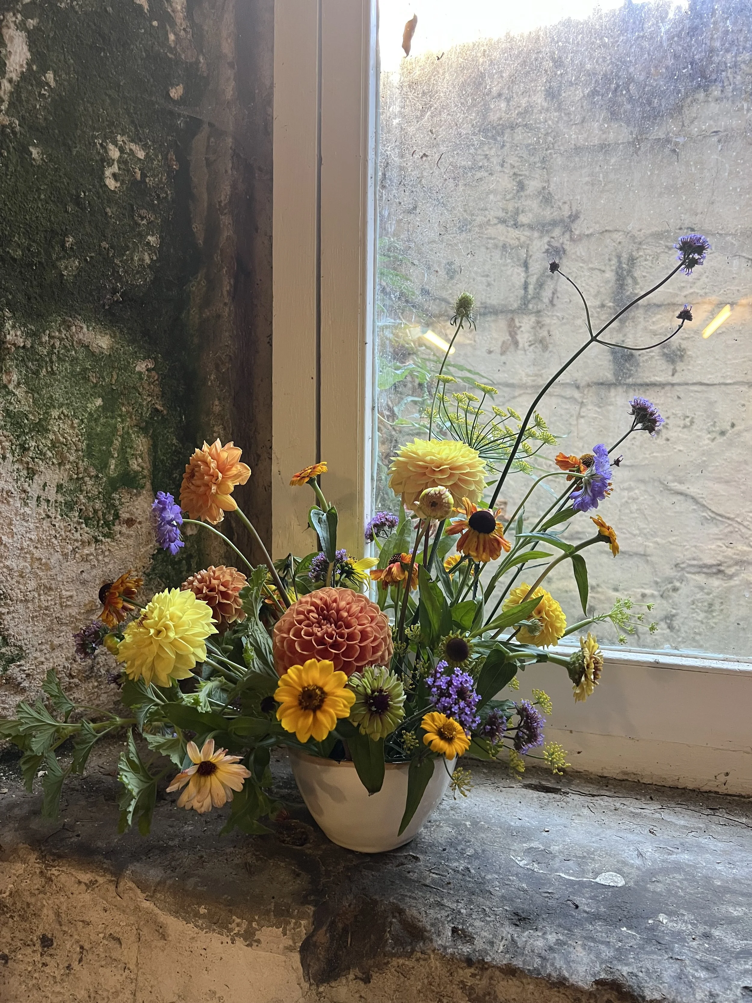 A bowl of flowers in a window recess, yellow, orange and purple british flowers