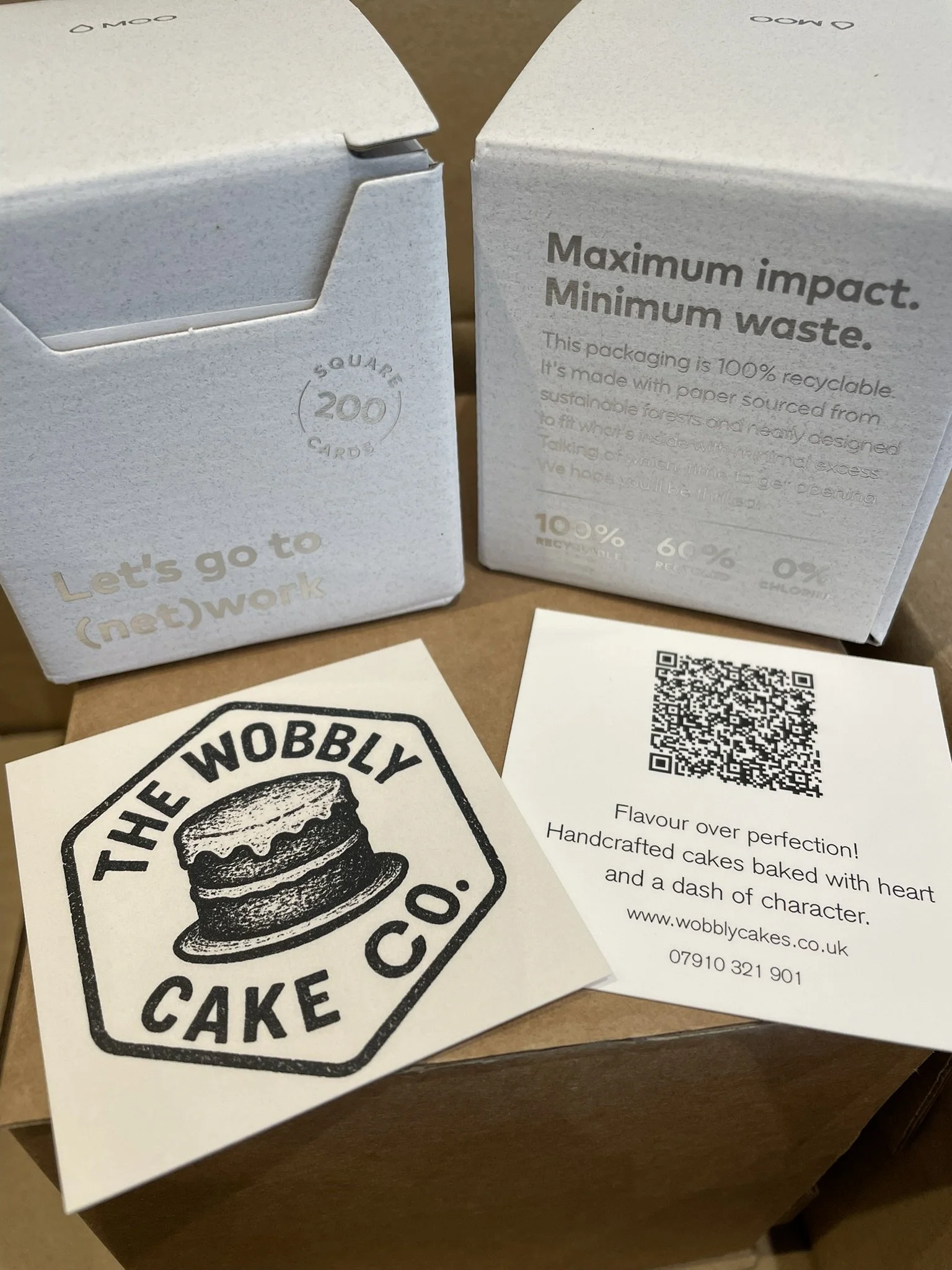 Two white cardboard boxes, one labeled 'Let's go to (net)work' and the other with recycling information about eco-friendly packaging. In front of the boxes is a business card with a logo depicting a cake and the text 'The Wobbly Cake Co.' along with a QR code and contact information.
