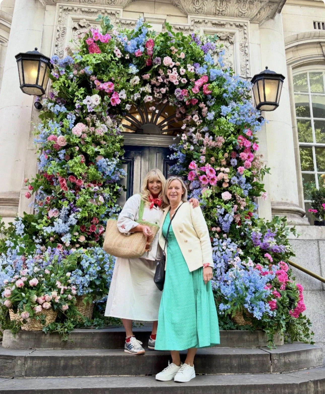 Two women standing on steps in front of a building entrance decorated with a large arch of pink, purple, blue, and white flowers. The women are smiling, one with a beige coat and a turquoise dress, the other with a white coat and cream skirt. They are holding bags and posing for the photo.