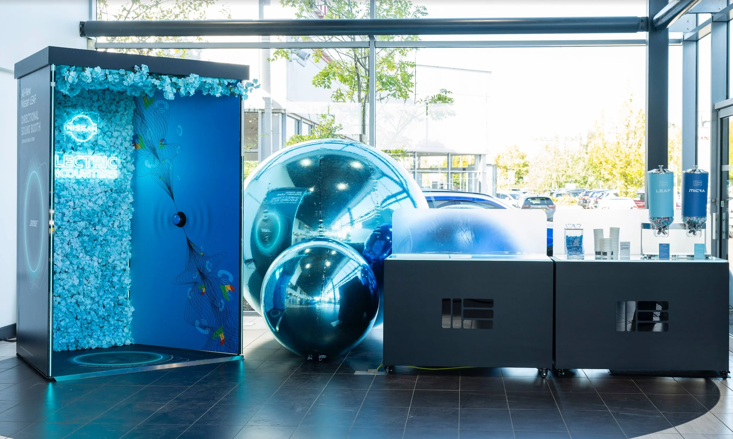 Nissan electric vehicle exhibit with a display of a large blue ball and a smaller metallic ball, promotional materials, and a blue flower wall, set inside a showroom with large glass windows and parking lot outside.
