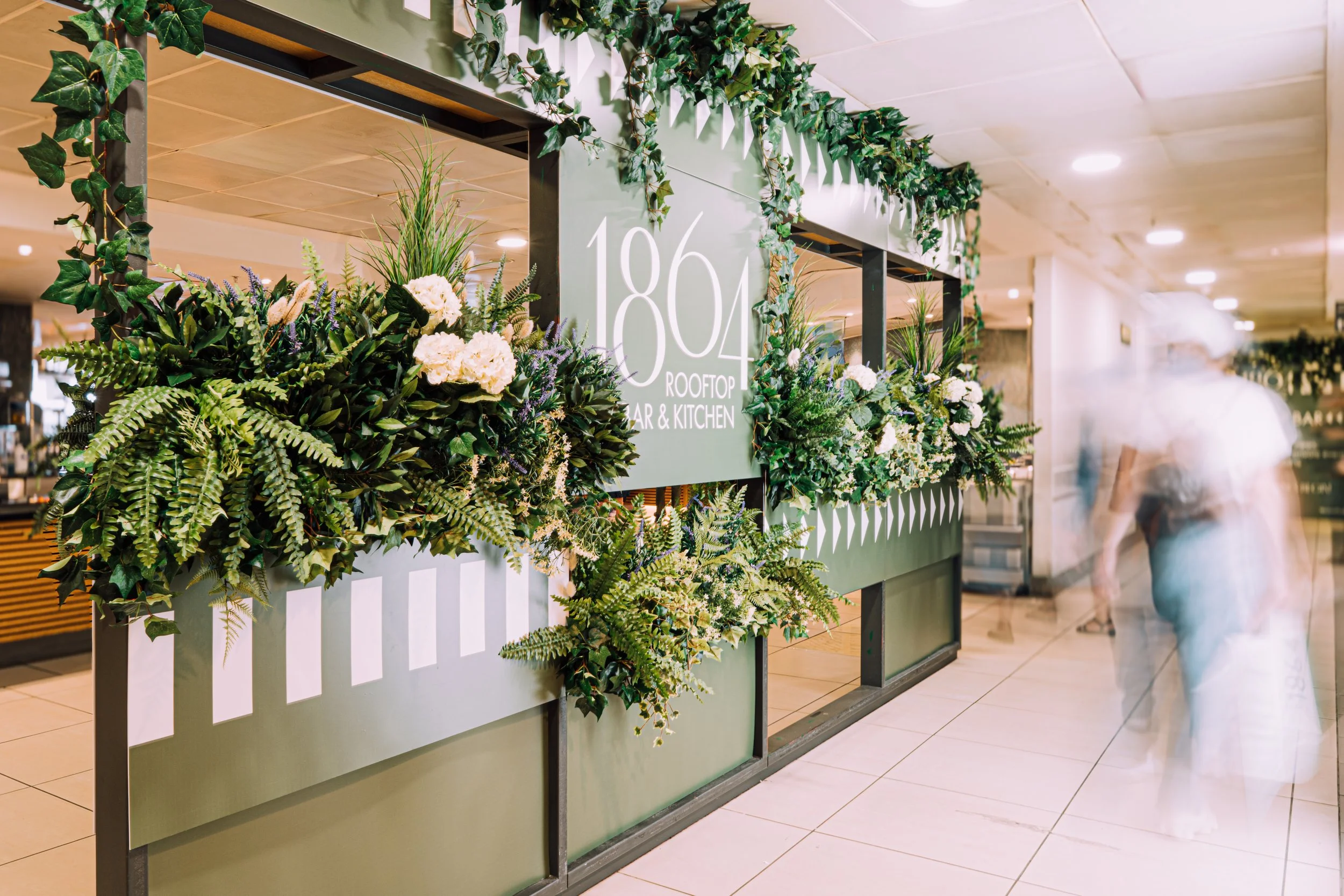 A floral display on a green stand with a sign that reads '1804 Rooftop Bar & Kitchen'. The display is decorated with various green plants and white flowers. There is an out-of-focus person in a white uniform walking past in an indoor setting.