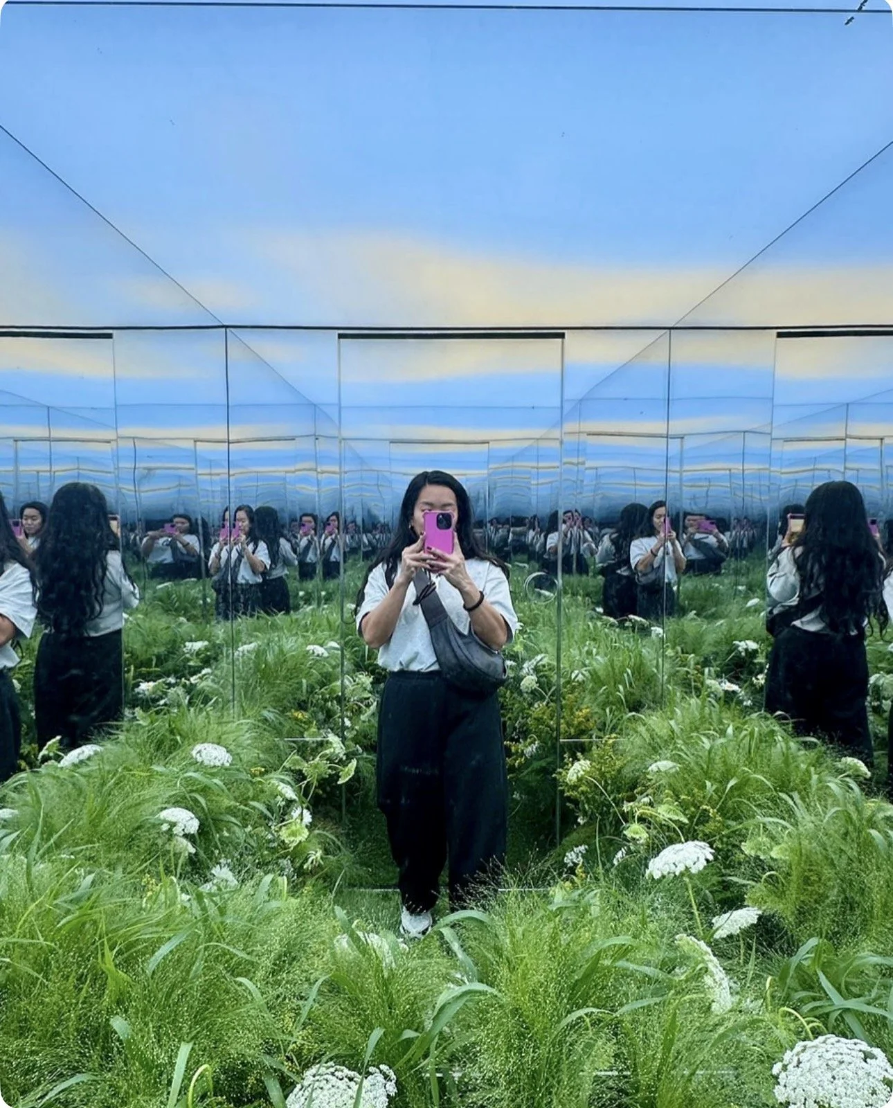 A person taking a photo of themselves with a pink phone in a room with mirrored walls, reflecting multiple images of themselves. The room is filled with green plants and white flowers, and the ceiling appears to be painted like a sky with clouds.