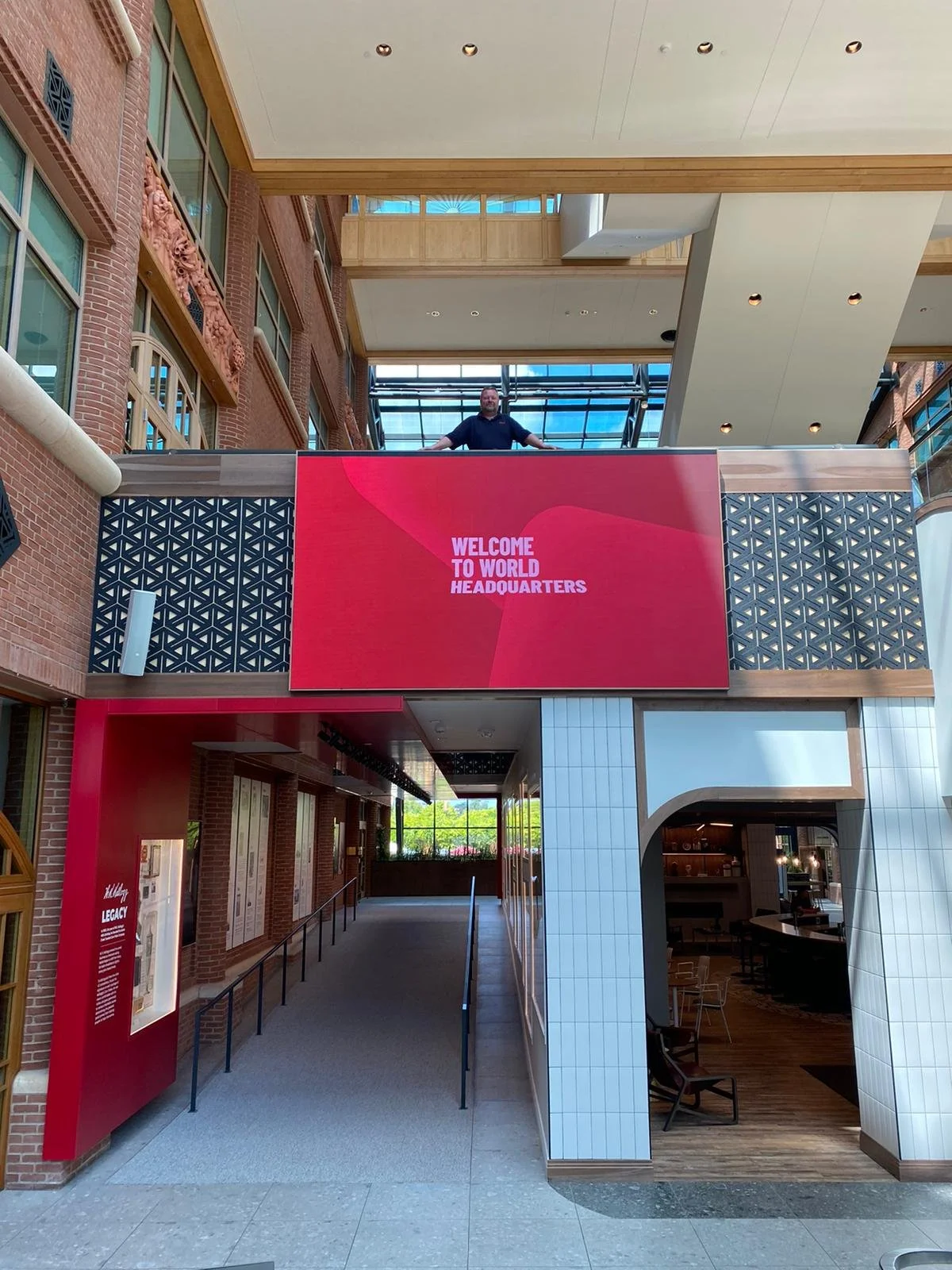 Interior of the Kellogg's headquarters with a large red digital screen displaying the message 'Welcome to World Headquarters'. A man is standing behind the screen on an upper level. The space has brick walls, large windows, and modern design elements.