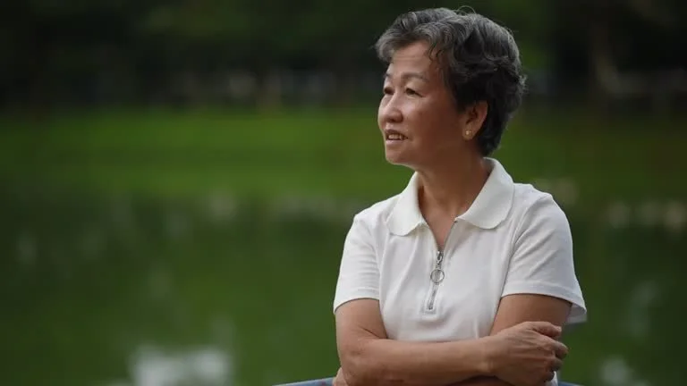 Older woman with short gray hair wearing a white polo shirt, standing outdoors near a body of water with green trees in the background.