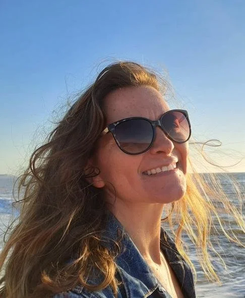 Woman with long hair wearing sunglasses and a denim jacket on a beach during sunset.
