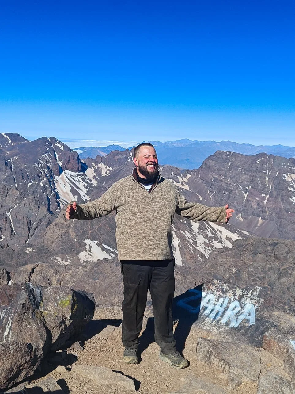 A person with short brown hair and a beard stands on top of a mountain with snow capped mountains in the background. He is smiling with arms outstretched. 