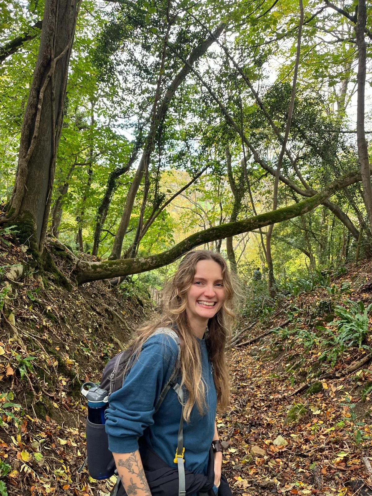 A person with long light brunette stands in the woods and hair and smiles at the camera