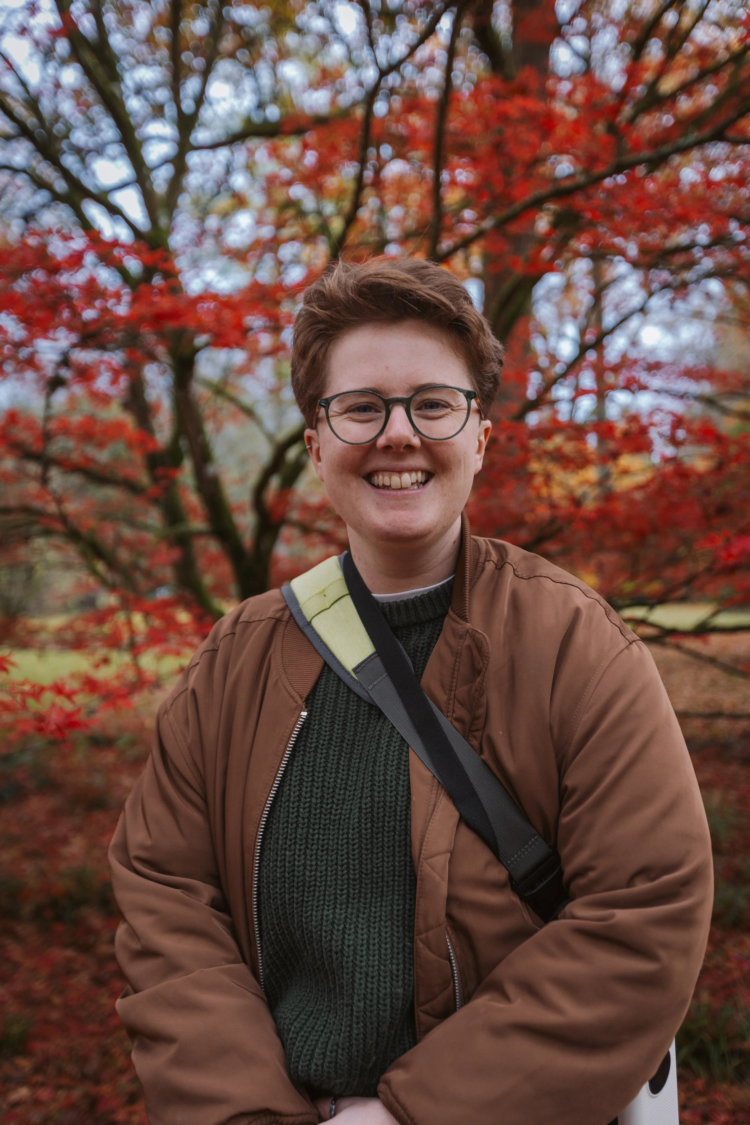 A white person with short brown hair smiles directly at the camera. They are outside with trees in the background.