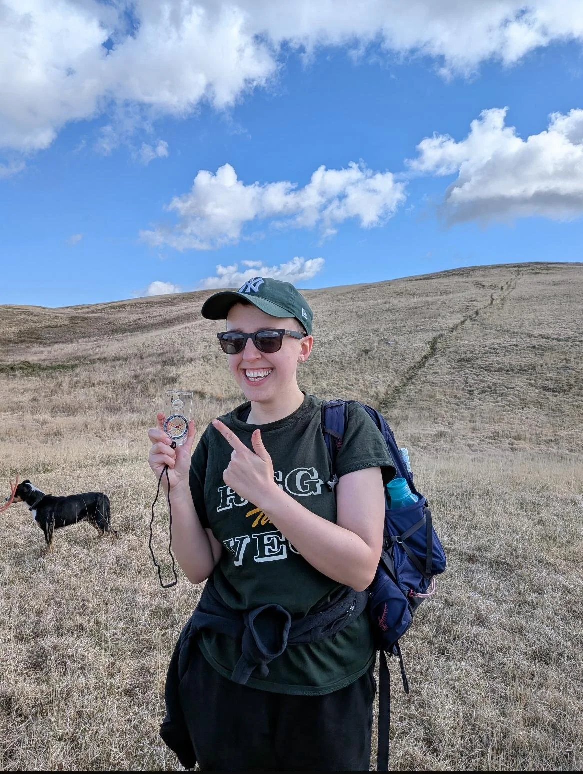 A person in a cap and sunglasses stands holding and pointing to a compass with a big smile on her face. She is in the countryside with a grassy hill behind her and blue skies.