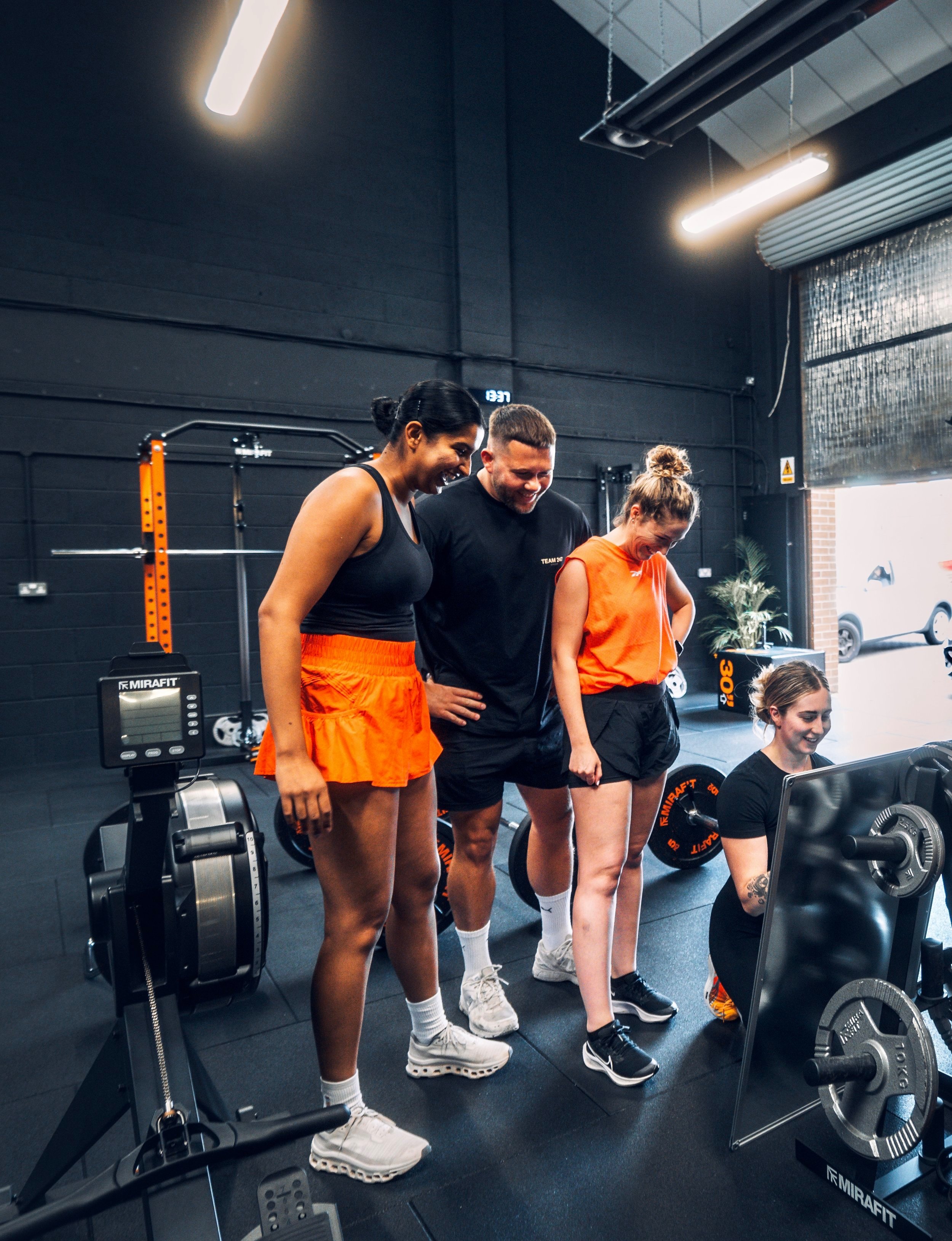 Four people with athletic clothing and bright orange shorts inside a gym, watching a woman lift weights with a smile.