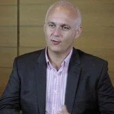 A bald man in a dark blazer and light-colored shirt sitting in front of a wooden background.