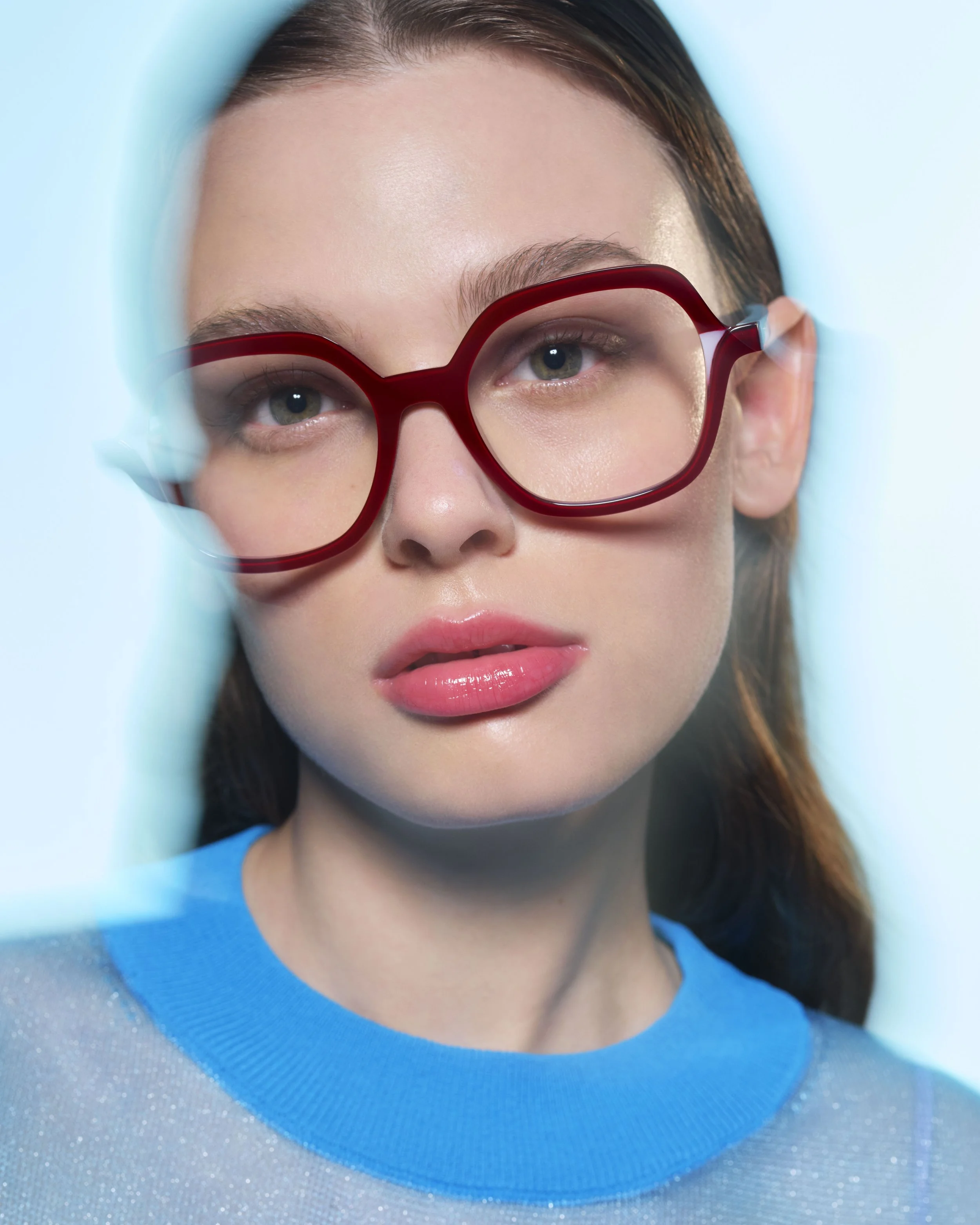 Une femme portant de grandes lunettes rouges, une chemise bleue et un maquillage naturel, regardant directement l'objectif.