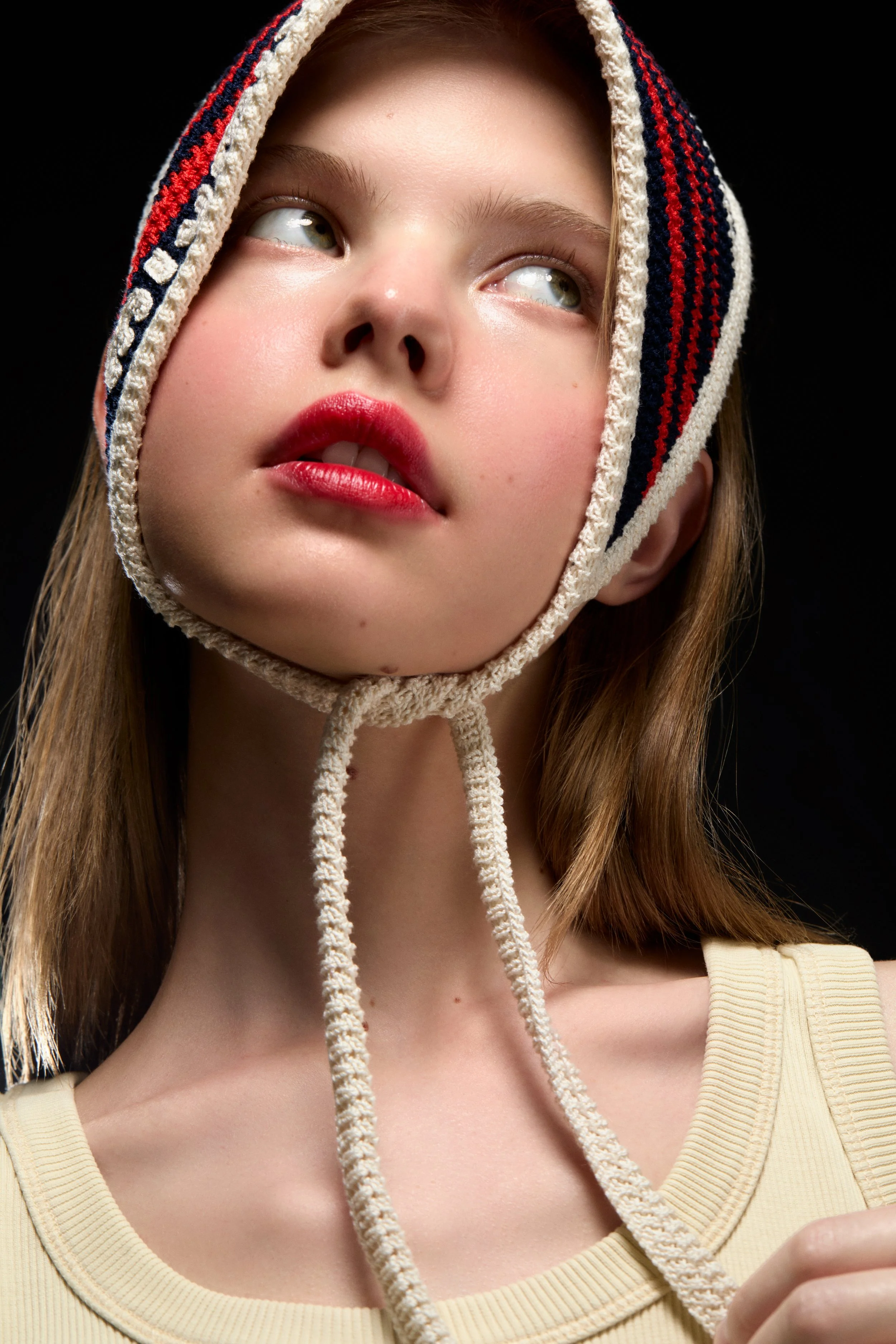 Jeune fille portant une chapka en tricot, avec des cheveux bruns, lèvres rouges, regardant vers le haut, sur fond noir.