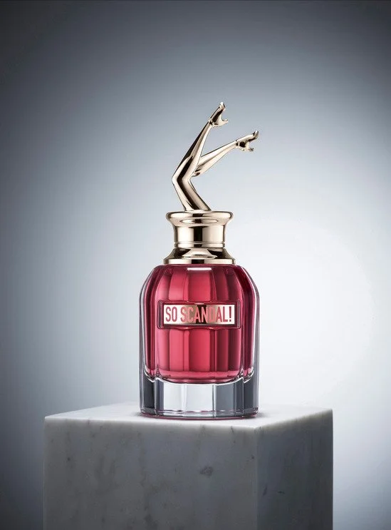 Un flacon de parfum rose avec un couvercle doré en forme de jambes féminines. Le flacon a une étiquette qui dit "SO SCANDAL!". Le flacon est placé sur une surface de marbre.