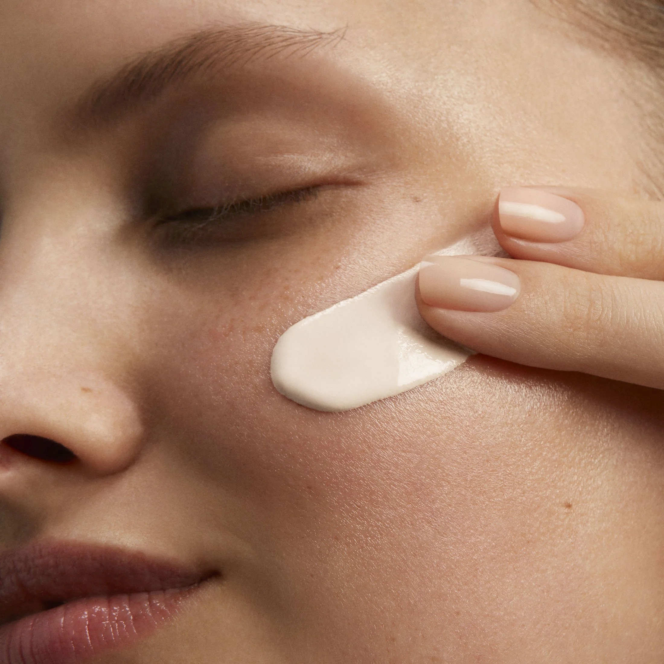 Une personne applique une crème blanche sur la joue avec deux doigts, la peau est claire et le visage est détendu.