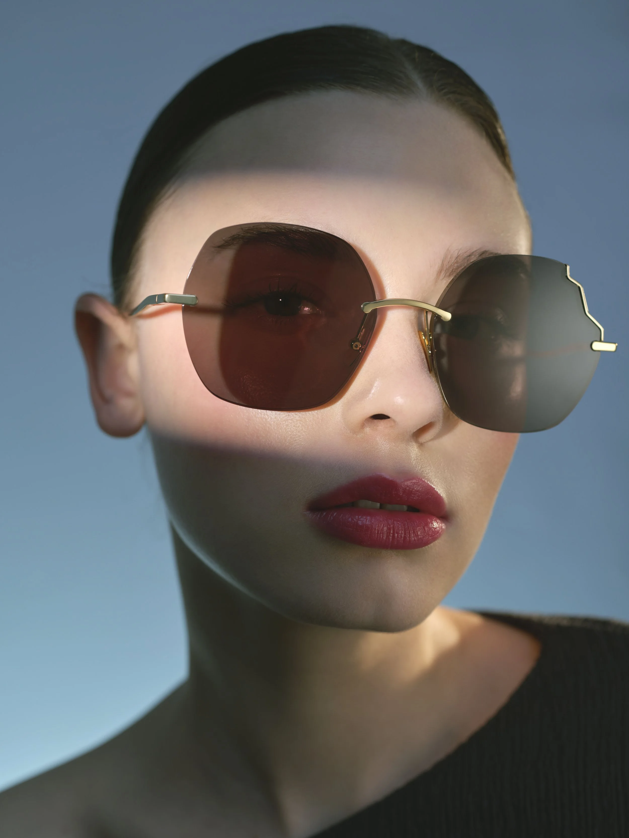 Une femme portant de grandes lunettes de soleil rondes et foncées, avec un maquillage sophistiqué, sur fond bleu clair.