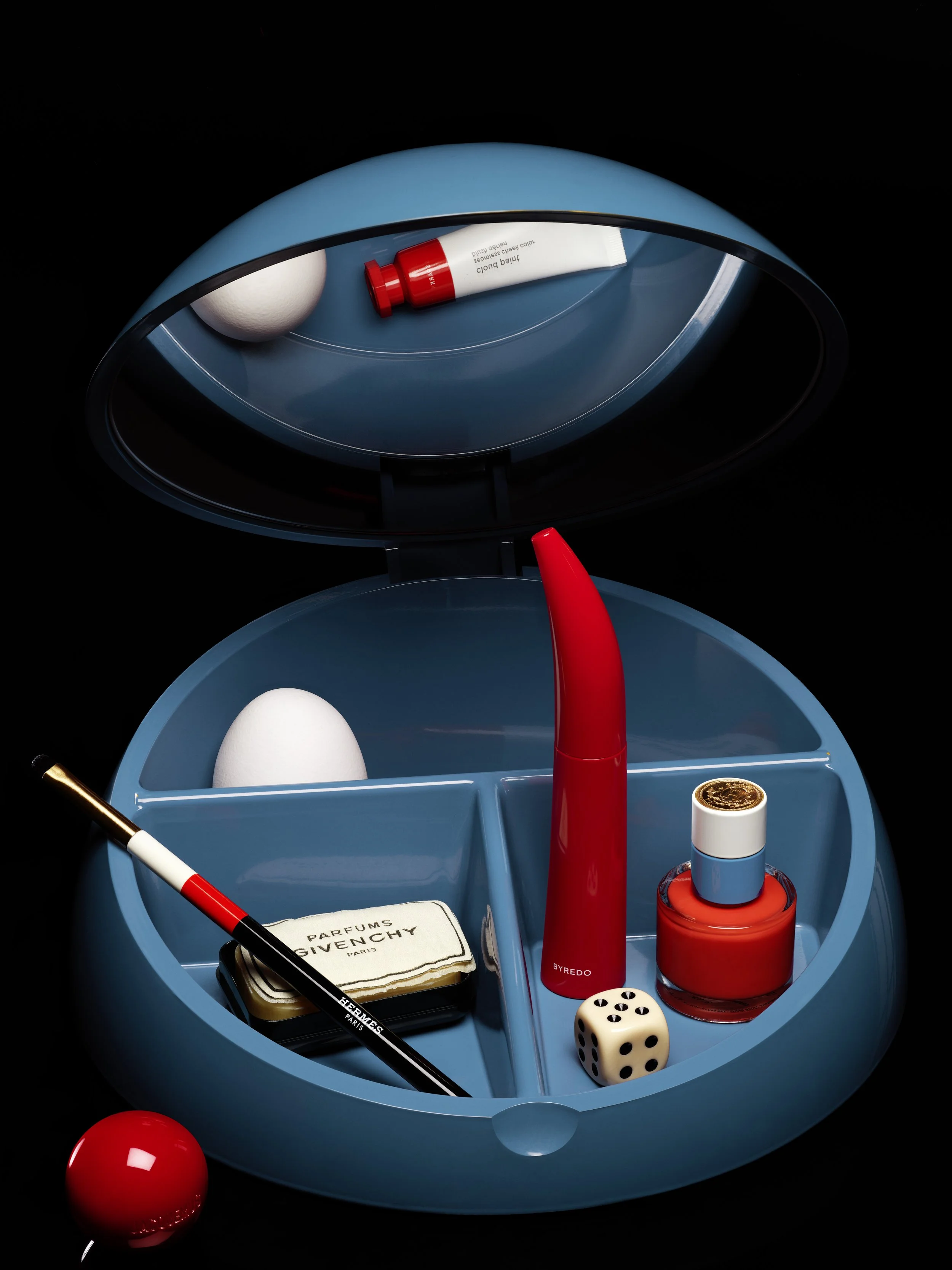 Une boîte à bijoux circulaire bleue contenant un œuf blanc, un flacon de parfum Givenchy, un pinceau, un spray rouge, un vernis à ongles rouge, un dé à jouer noir et blanc, une boule rouge à l'extérieur