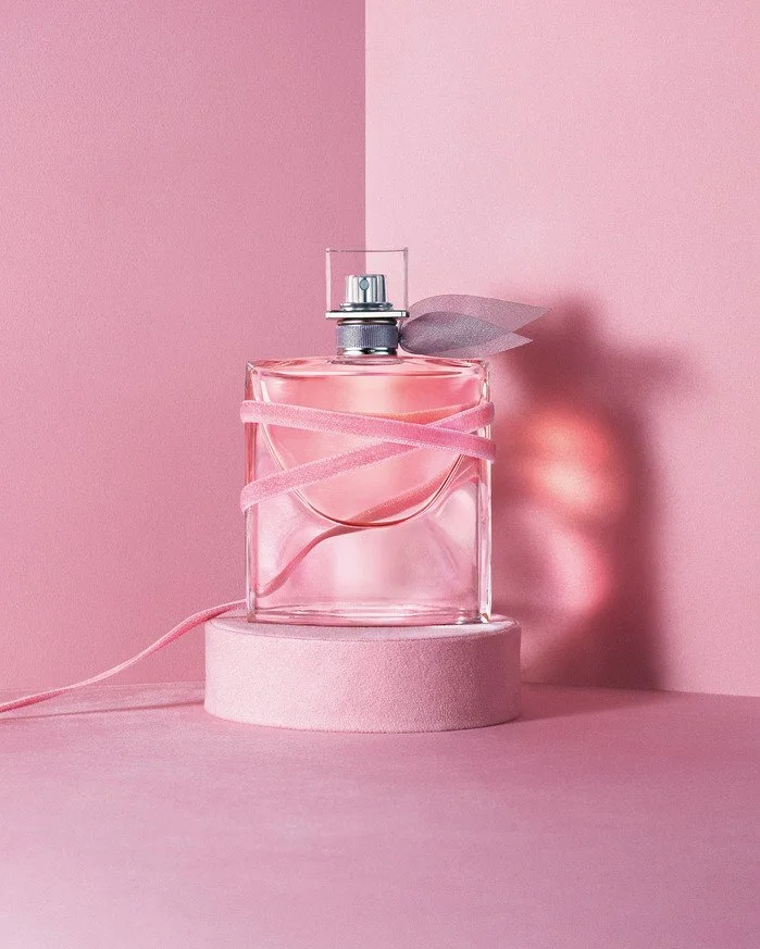 Flacon de parfum transparent avec une garniture rose, posé sur un support rond en mousse rose, avec un fond et sol rose