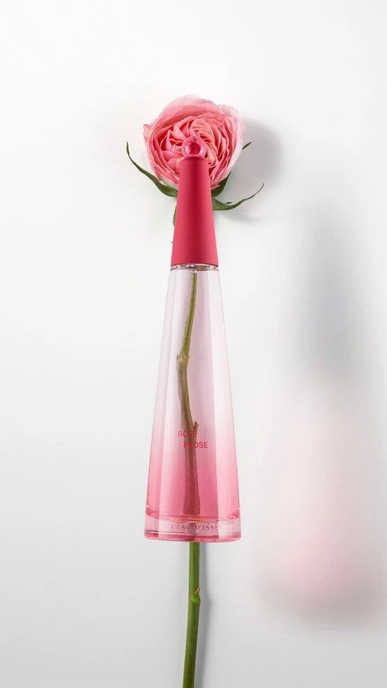 Un flacon de parfum rose placé sur une tige de rose, la tige forme le corps du flacon. La fleur de rose est derrière le flacon, créant une composition esthétique.