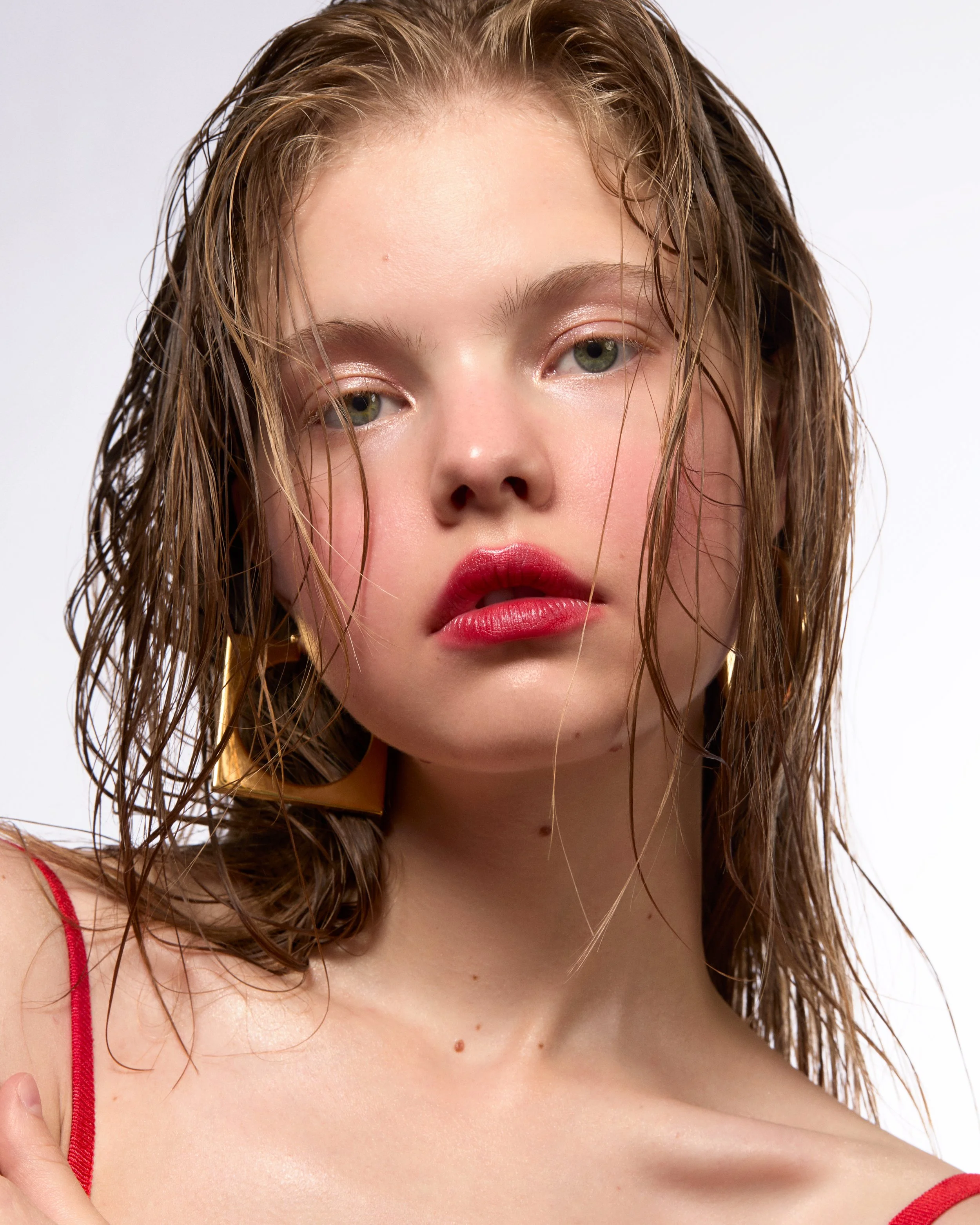 Jeune femme aux cheveux mouillés, maquillage rosé et lèvres rouges, portant une robe à bretelles rouges, portant des boucles d'oreilles en forme de triangle dorées.