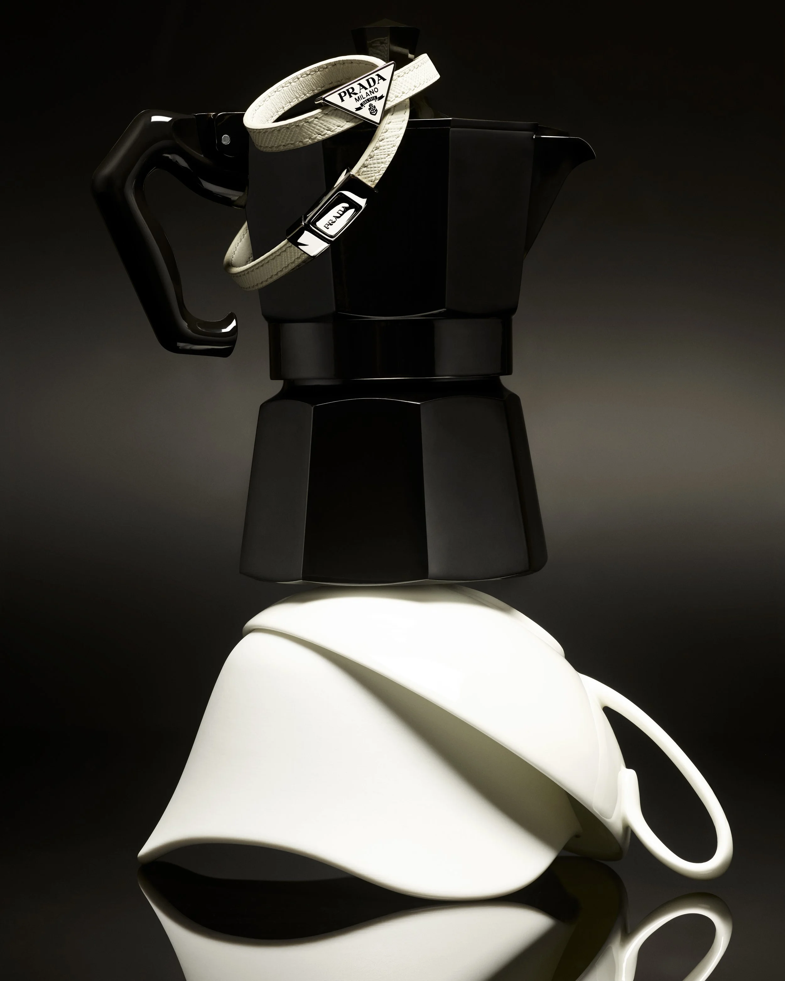 Une cafetière moka noire avec un porte-filtre posé dessus, décorée avec un bracelet blanc Prada et livré dans un fond sombre.