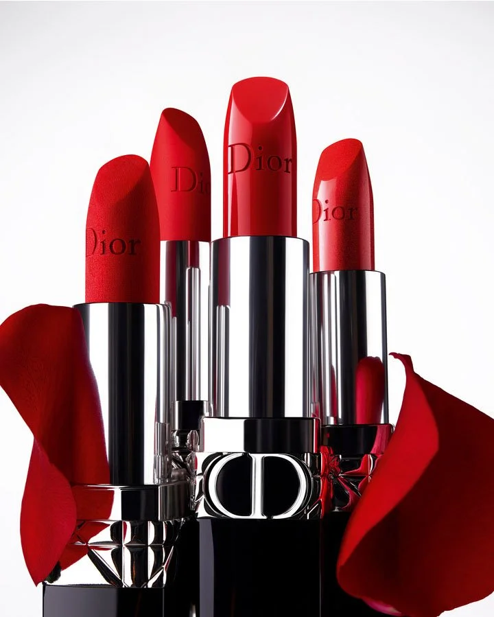 Quatre rouges à lèvres Dior avec des pétales de rose rouge autour, sur un fond blanc.