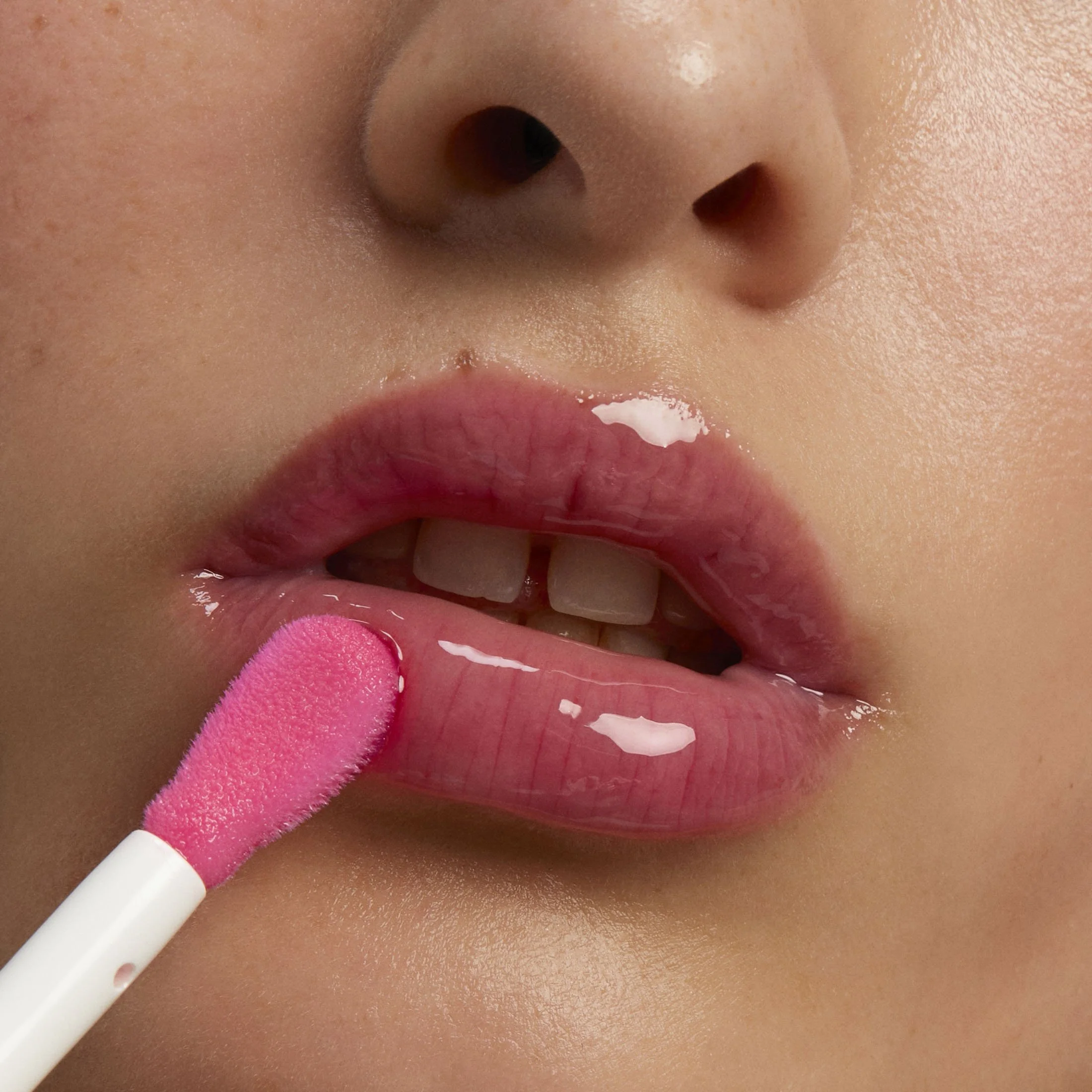 Close-up d'une personne appliquant du gloss rose sur ses lèvres pulpeuses, avec un nez visible en arrière-plan.