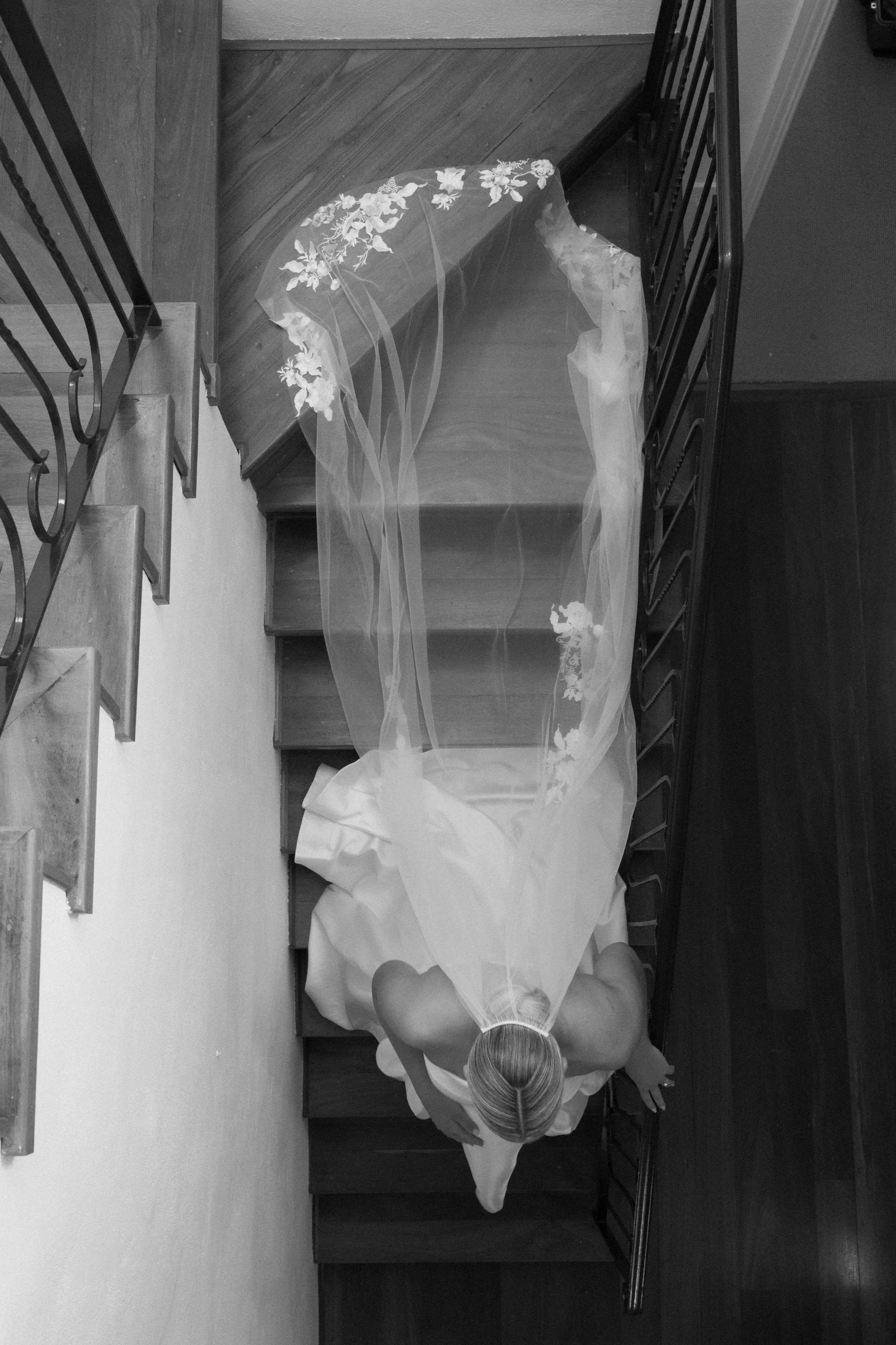 A woman in a wedding dress standing at the top of a staircase, viewed from above in black and white.