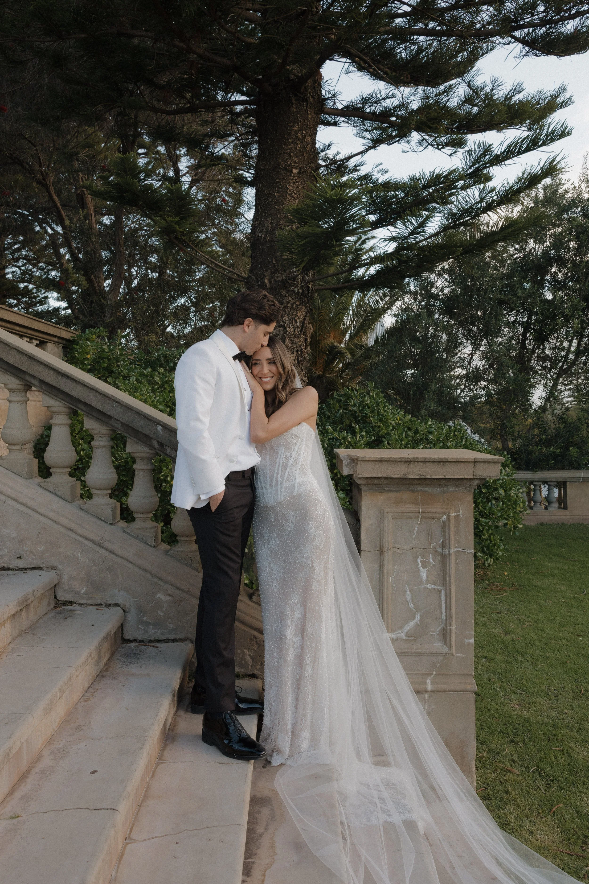 A bride and groom are standing on stone stairs outdoors, embracing each other. The groom is wearing a white tuxedo jacket, black pants, and a bow tie, while the bride is in a white wedding gown with a long veil. There are trees and a stone railing in