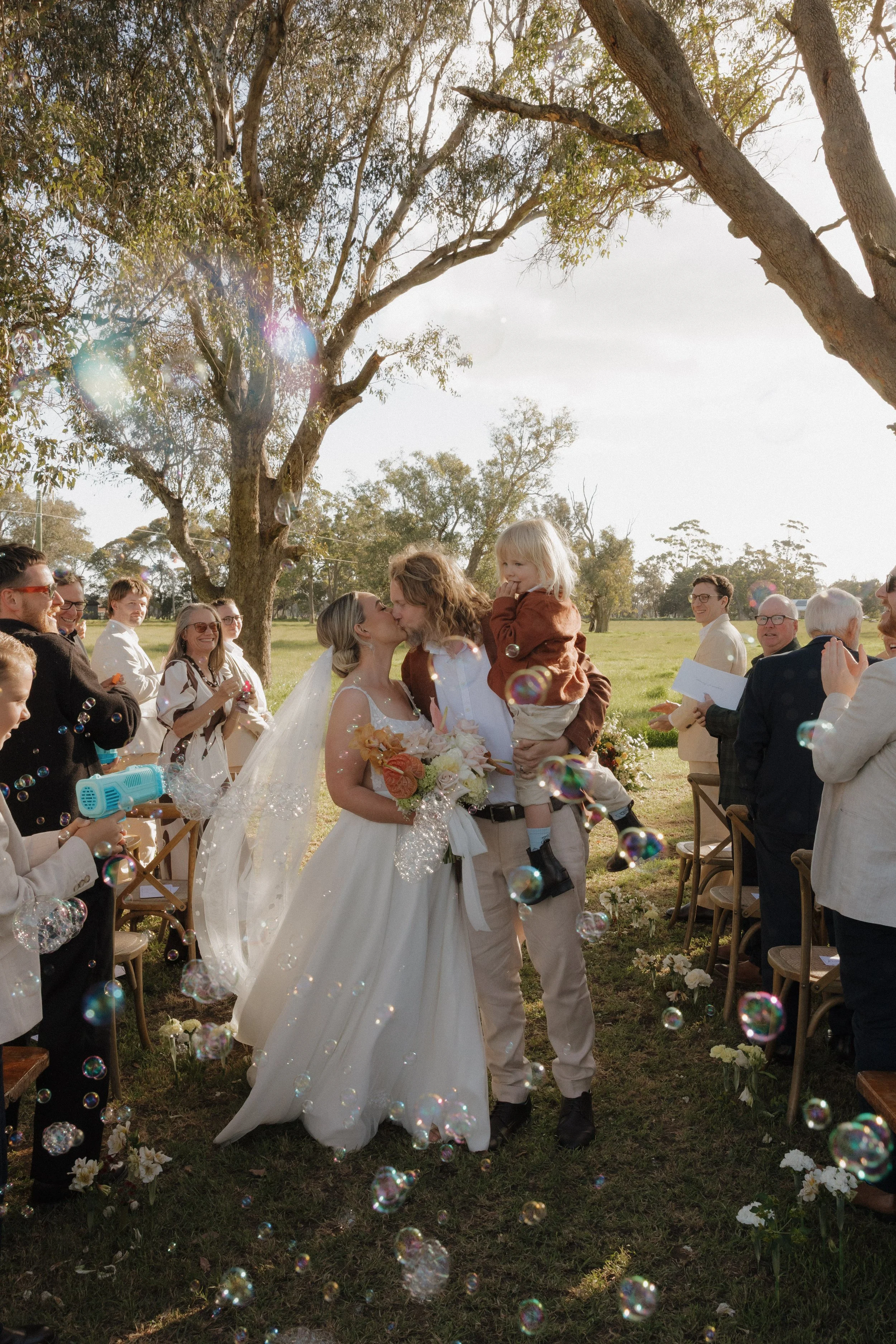 A bride and groom share a kiss during their outdoor wedding ceremony, surrounded by guests, trees, and bubbles in the air.