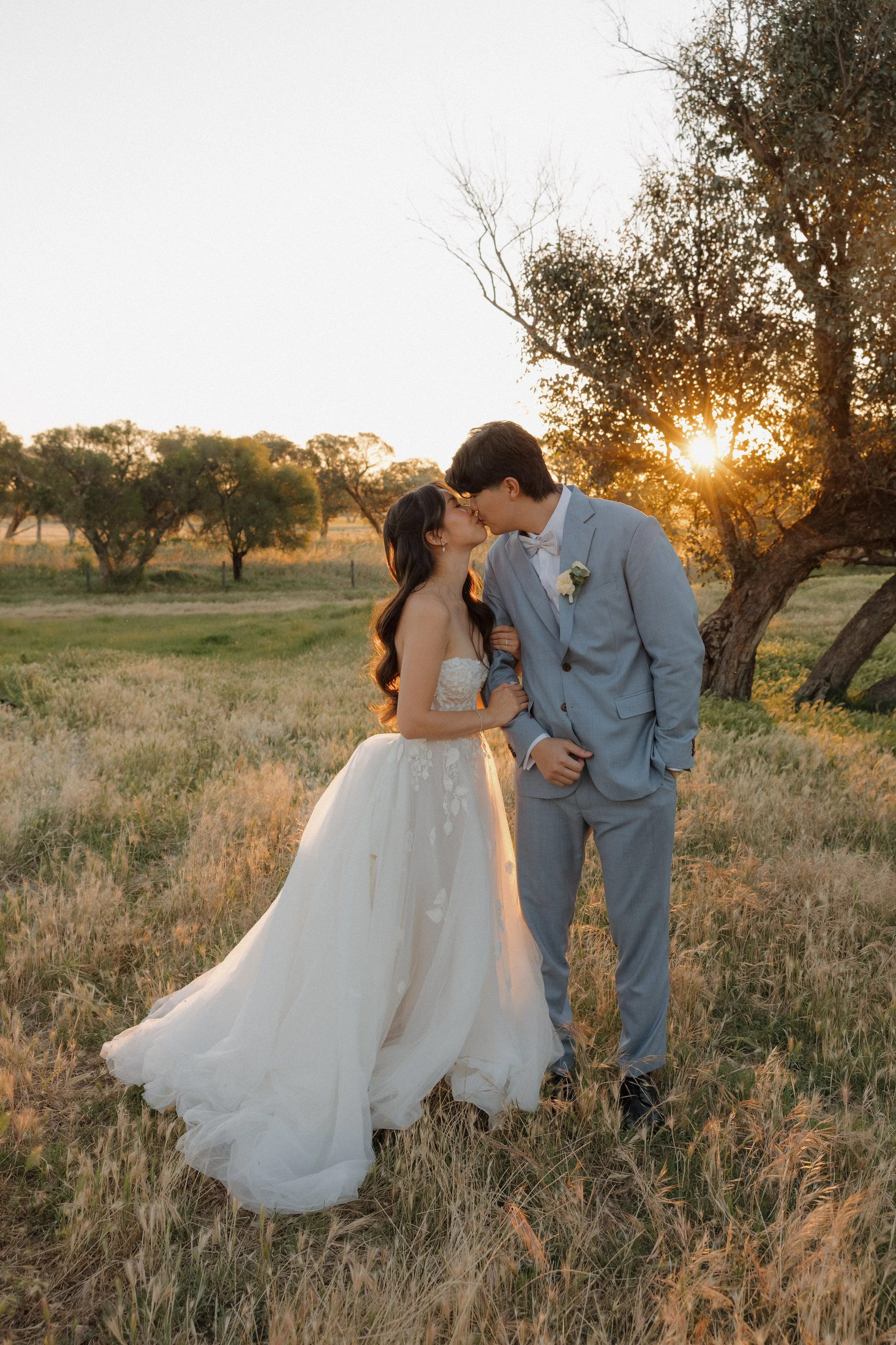 A bride and groom kiss outdoors at sunset in a grassy field with trees in the background.