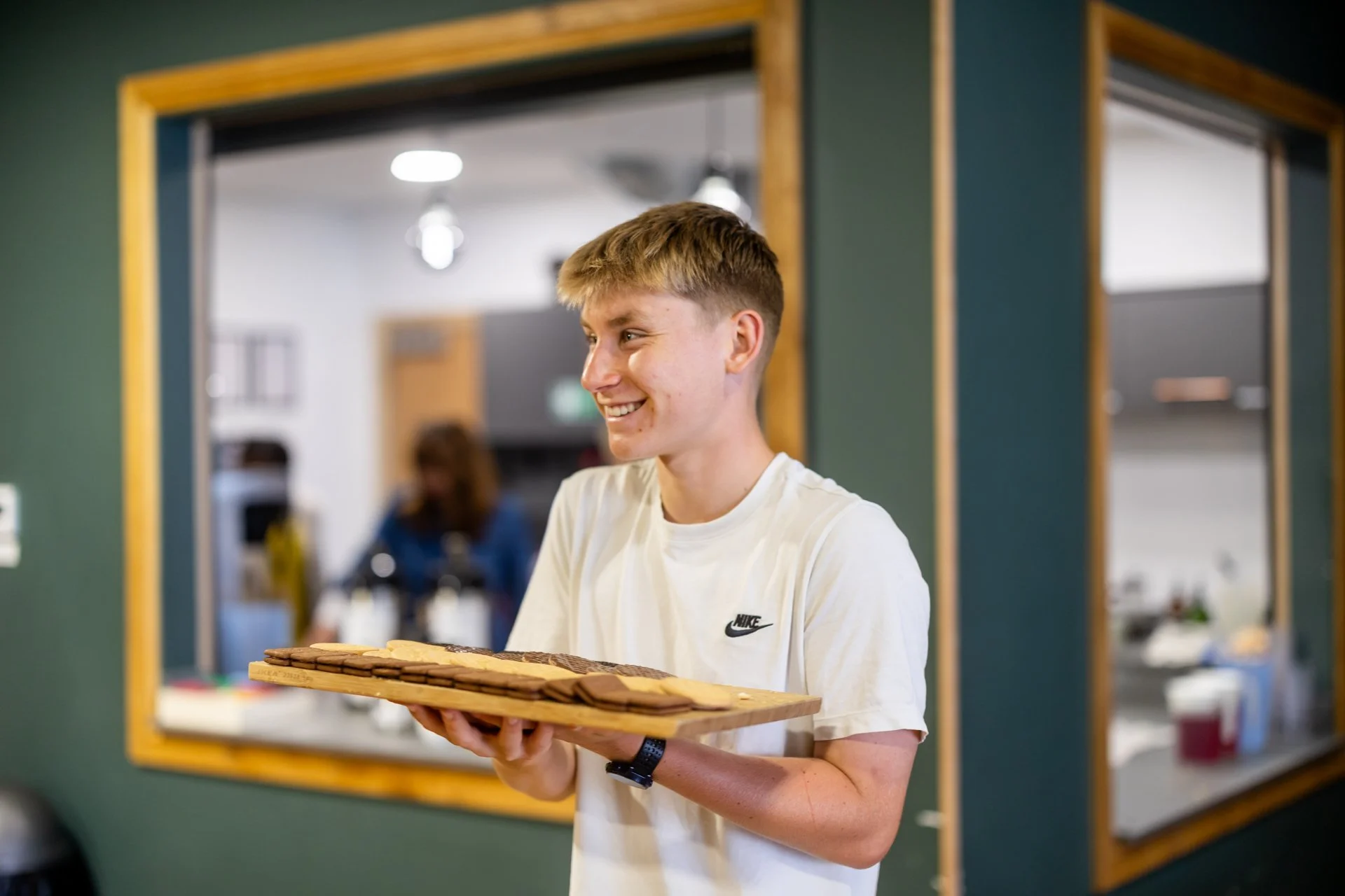 A young man wearing a white Nike t-shirt is smiling and holding a large tray of assorted cookies. He is standing in front of a green wall with a window looking into a kitchen or common area.