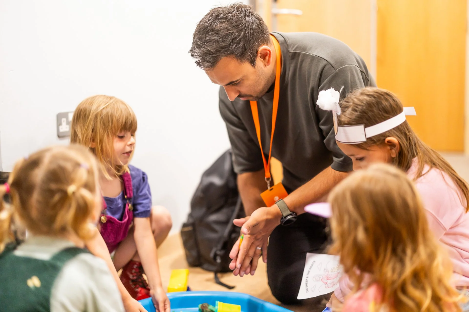 A man with short dark hair and wearing a dark gray shirt and an orange lanyard is kneeling on the floor, engaging with four young girls with red and blonde hair. The girls are gathered around a blue container with various colorful objects inside, in