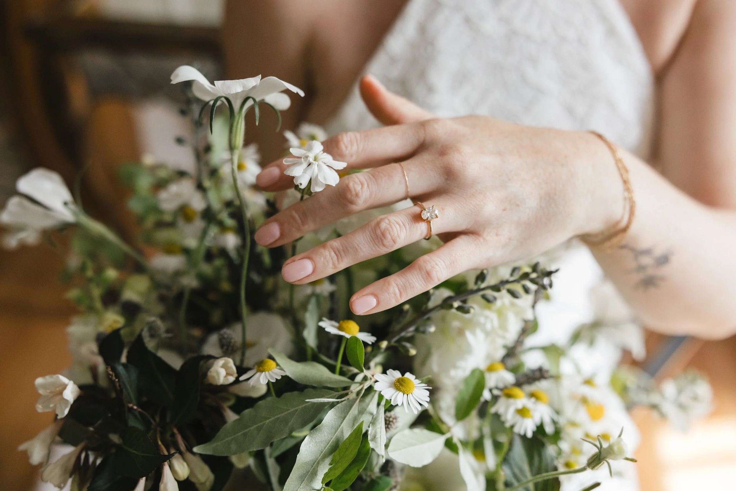 Flroal design in Connecticut shows a Close-up of a Bride's hand with a wedding ring touching her white bridal bouquet, with visible tattoos and a delicate bracelet.
